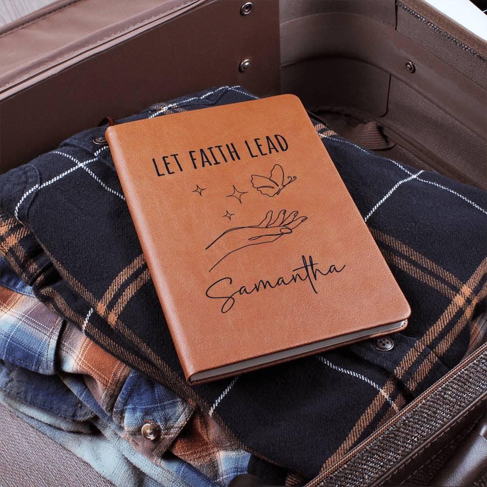 The Personalized Leather Prayer Journal for Women with "Let Faith Lead" lies stylishly on plaid shirts in a suitcase, making it an ideal Christian gift.