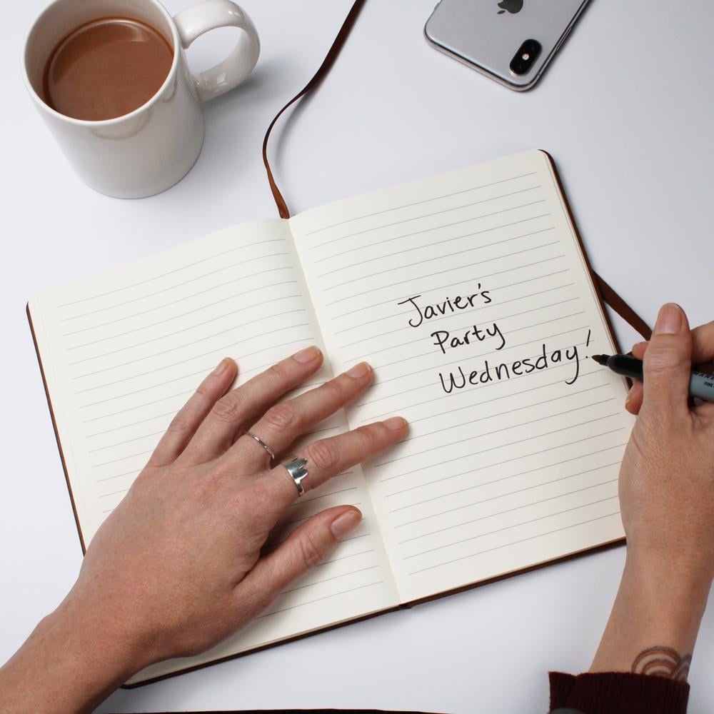 A person writes "Javier's Party Wednesday!" in a lined notebook with a black marker, next to a hot chocolate, iPhone, and a Personalized Leather Cycling Journal For Men—an ideal gift for road bikers and cyclists. Rings and a tattoo decorate their wrist.