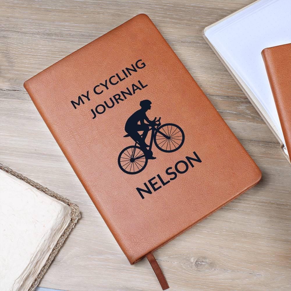 A brown Personalized Leather Cycling Journal For Men, custom name notebook with ribbon bookmark and black cyclist silhouette on the cover. A perfect gift for road bikers and cyclists, shown on a wooden surface beside books and a placemat.