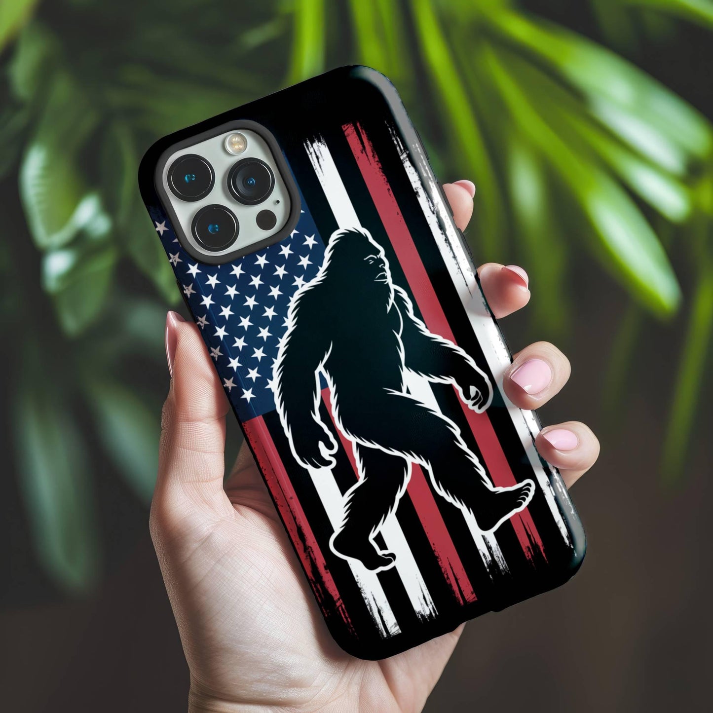 A hand holds the Golden Value SG's Bigfoot Silhouette Phone Case features a Bigfoot image set against a distressed American flag, complete with stars on a blue field and red and white stripes. Green foliage is softly blurred in the background.