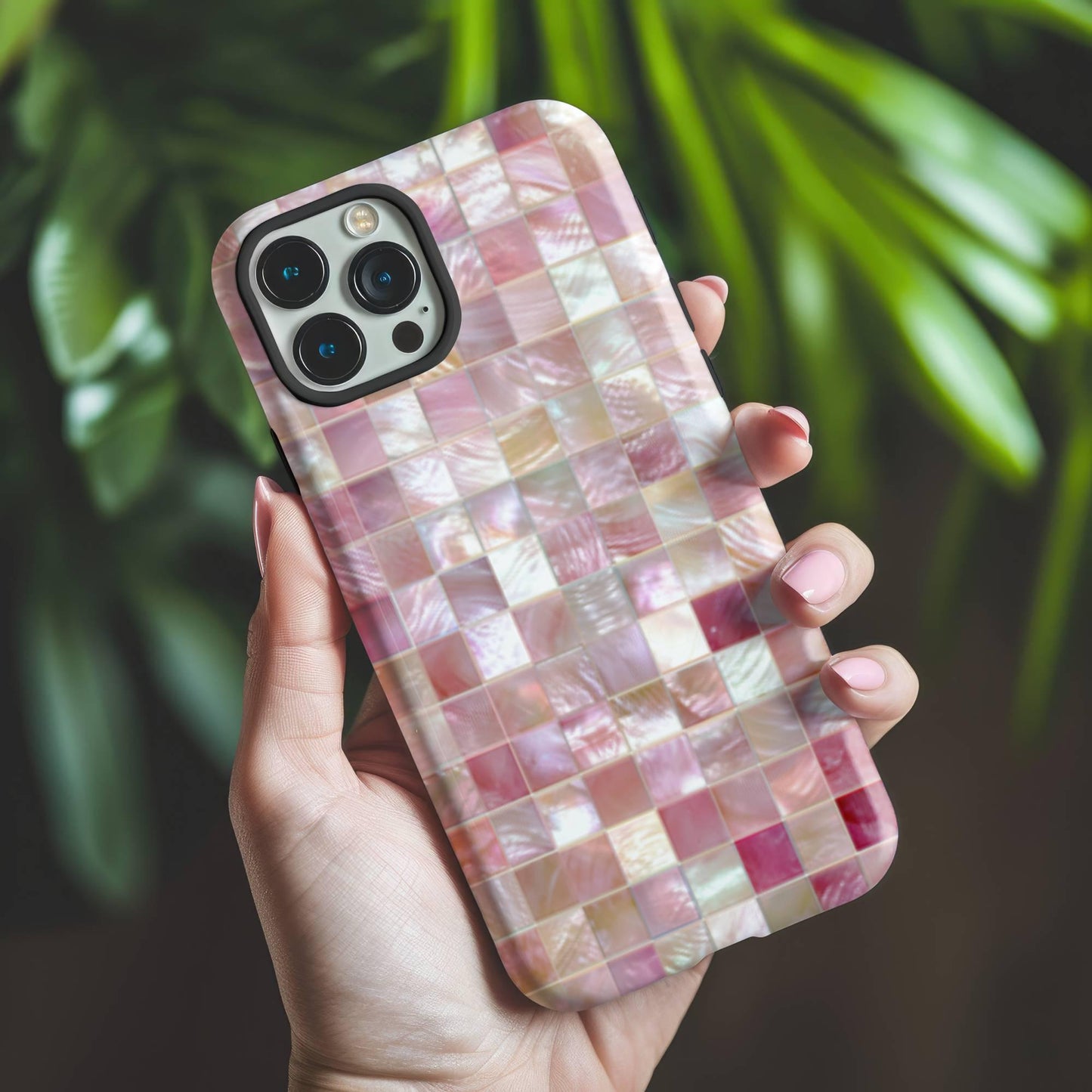 A hand holds the Mother of Pearl Pink Mosaic Phone Case by Golden Value SG featuring a mosaic pattern of shimmering, iridescent tiles in light pink, white, and pastel hues, creating a visual texture with mother-of-pearl effects. Green foliage is softly blurred in the background.