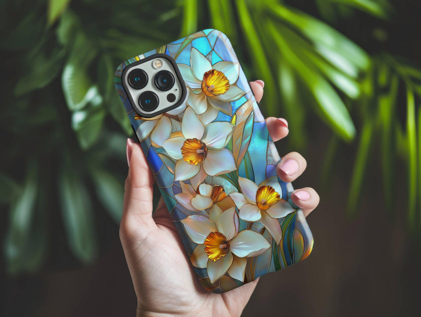 A hand holds a Stained Glass Daffodil Phone Case by Golden Value SG featuring elegant white daffodils with vibrant yellow centers and green leaves against a mosaic-style blue and teal background. This floral design captures the delicate beauty of spring flowers in a colorful, art-inspired pattern, perfect for adding a touch of artistic charm to your device. Green foliage is softly blurred in the background.