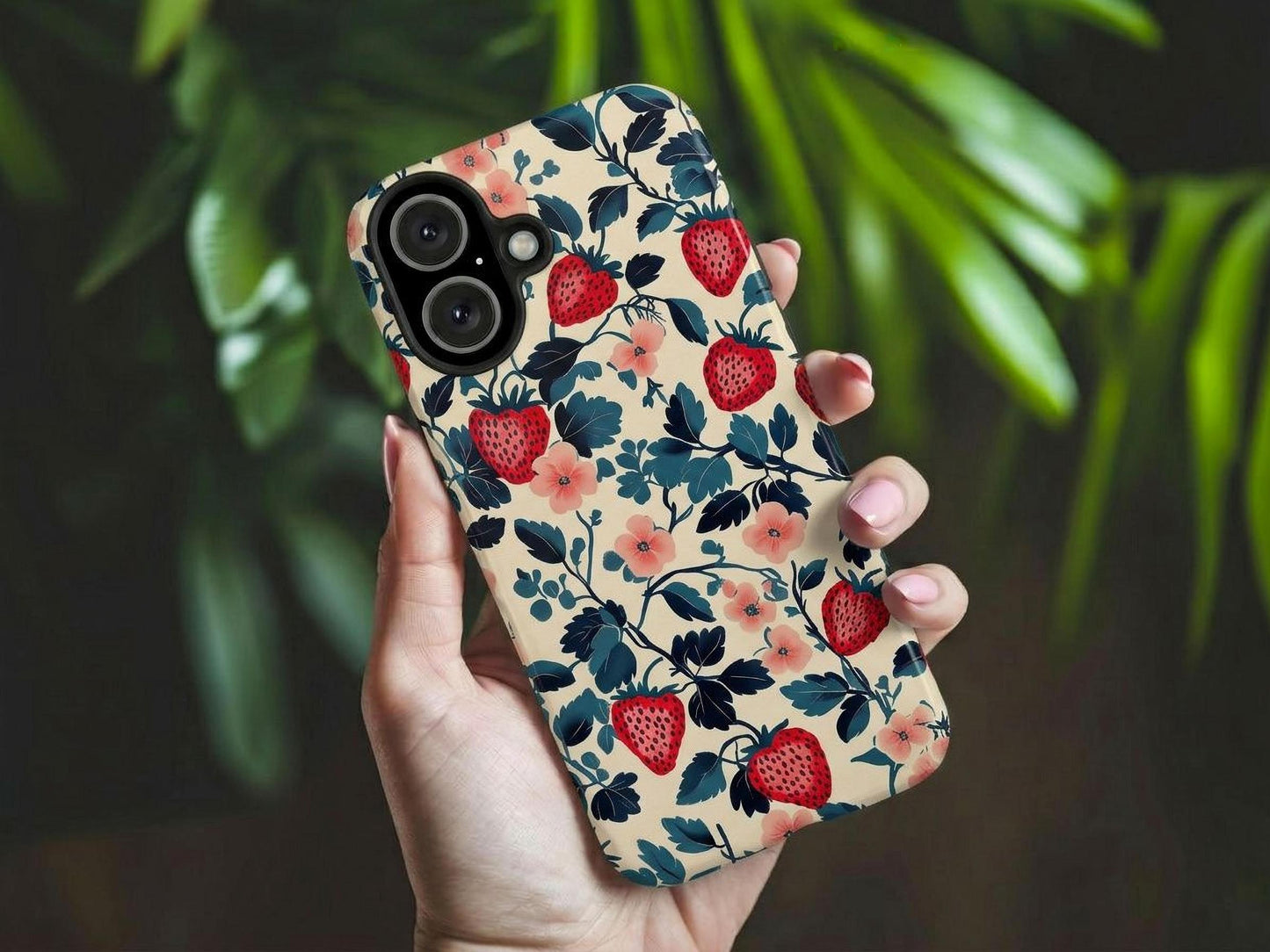 A hand with light pink manicured nails holds the Golden Value SG Strawberry Phone Case, featuring red strawberries, navy blue leaves, and pink flowers on beige. Green leaves blur in the background.
