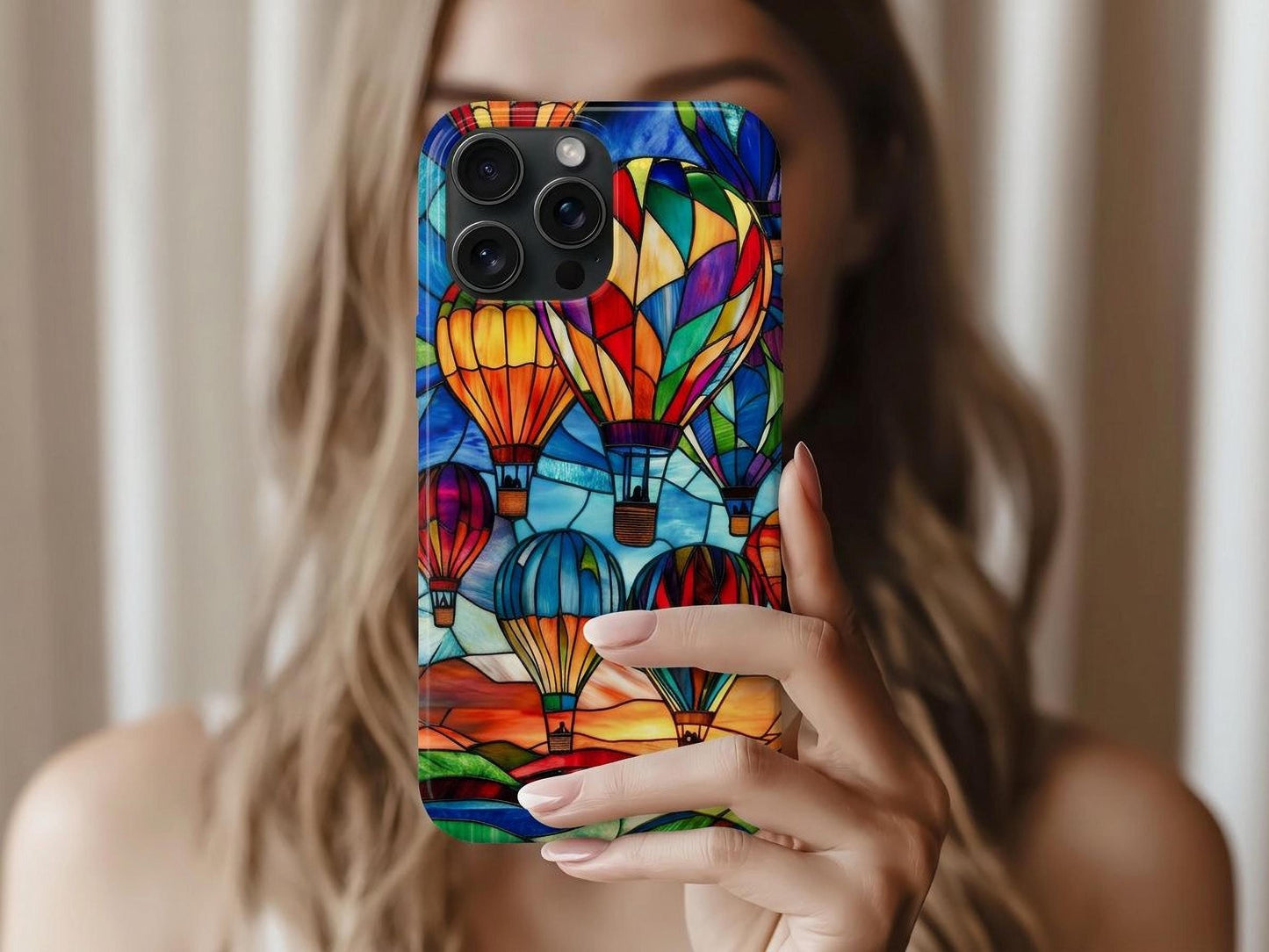 A woman with light brown hair holds up a Golden Value SG's Hot Air Balloon Phone Case features stained glass artwork of vibrant hot air balloons in red, blue, green, and yellow against a sunset landscape with a sky turning from deep blue to orange.