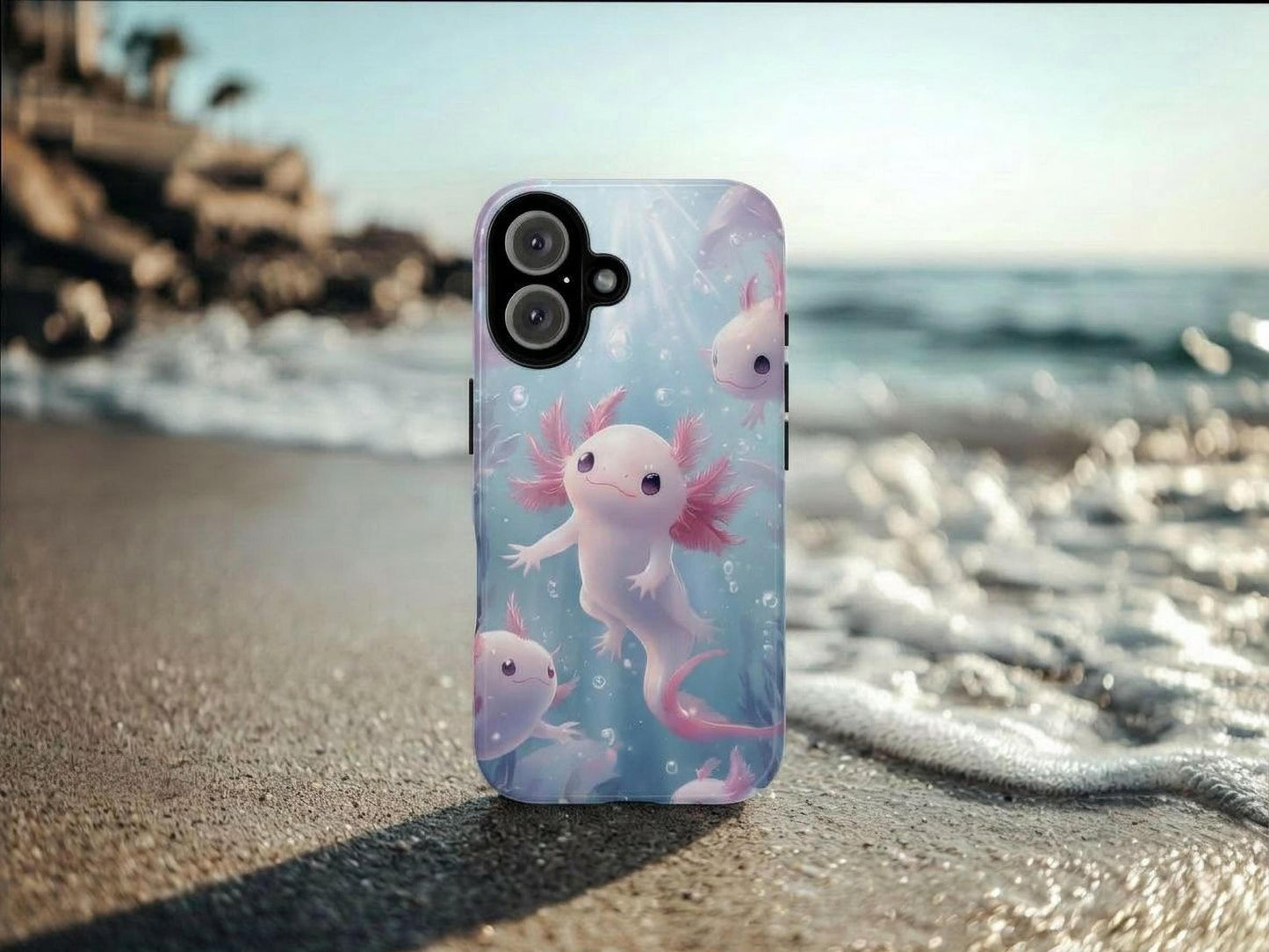 A Golden Value SG Cute Axolotl Salamander Phone Case, showing smiling pink axolotls swimming in blue water, rests on wet beach sand by the shore with gentle waves and blurred ocean rocks beneath a clear sky.