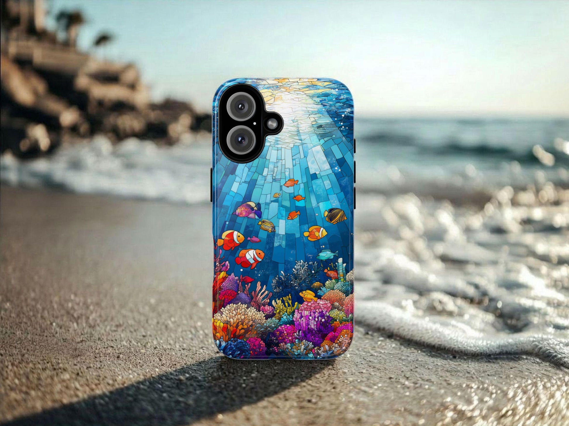 The Golden Value SG Stained Glass Ocean Phone Case, featuring vibrant fish, coral, and sunbeams, stands upright on wet sand by the ocean with gentle waves and a blurred shoreline in the background.