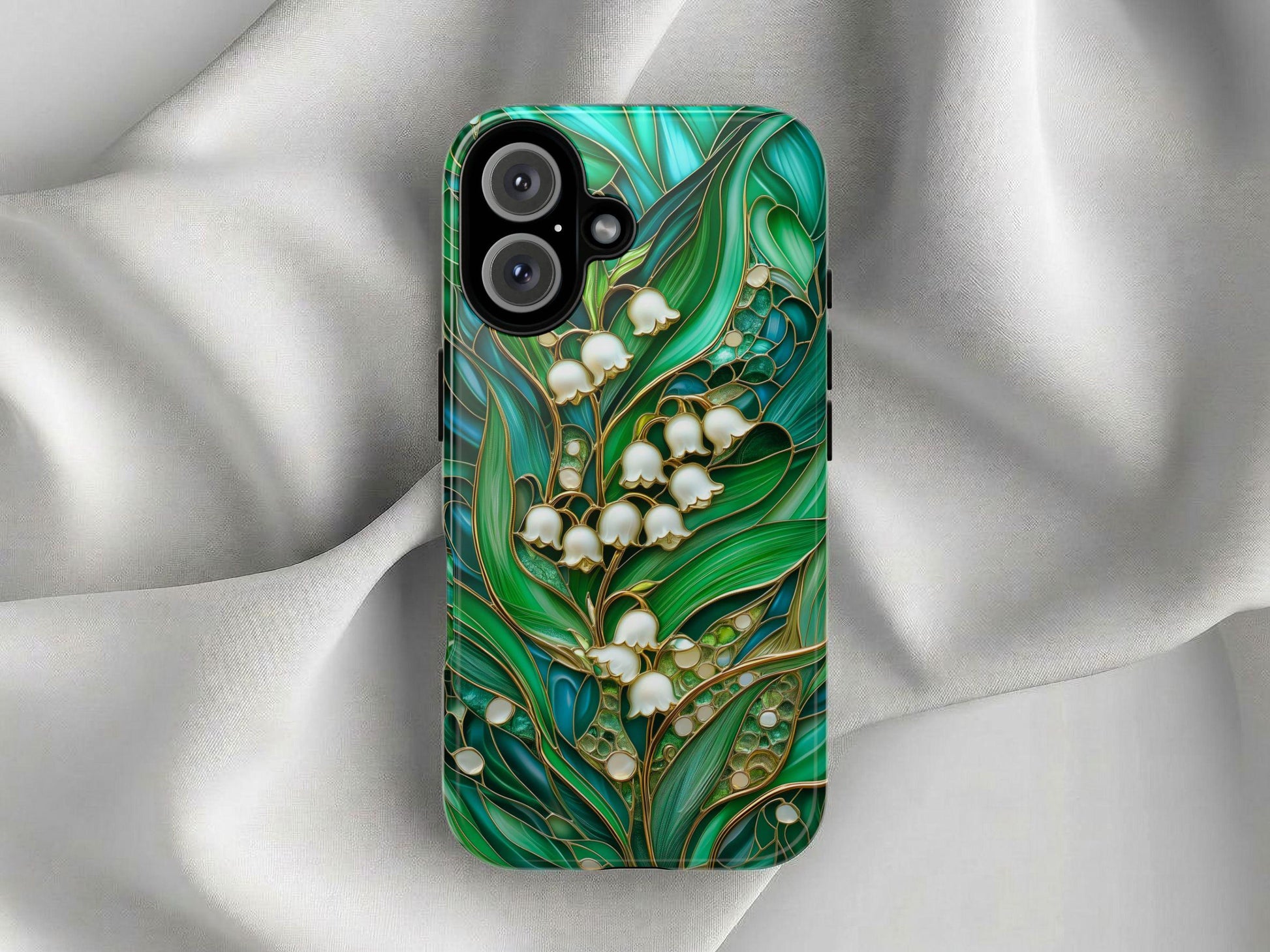 A Stained Glass Lily of the Valley Phone Case by Golden Value SG, with a glossy finish and vibrant green leaves and white lily of the valley pattern, rests on soft, silver-gray fabric.