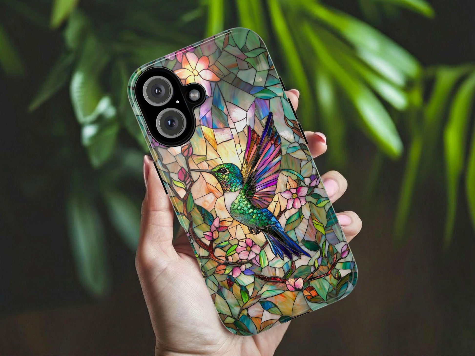 A hand holds a Golden Value SG's faux stained glass Hummingbird Phone Case showcases a colorful hummingbird surrounded by pink flowers and green leaves, set against a vibrant, multi-colored background. Green foliage is softly blurred in the background.