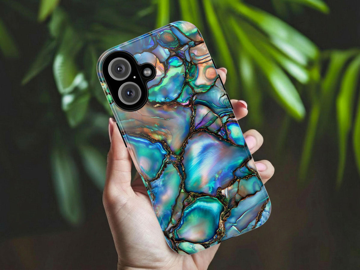 A lady's hand is holding an iphone Abalone Shell Design Phone Case featuring the iridescent beauty of the case delivers a mesmerizing play of colors that mimic the captivating allure of gemstones. Against the background of some green plants.