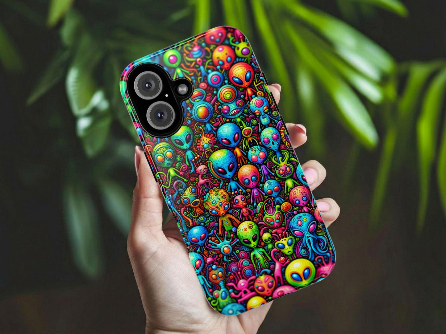 A hand holds the Whimsical Alien Phone Case by Golden Value SG features a vibrant collage of cartoon-style aliens and space creatures with unique shapes, big eyes, and vivid blue, pink, green, orange, and yellow hues densely packed together. Green foliage is softly blurred in the background.