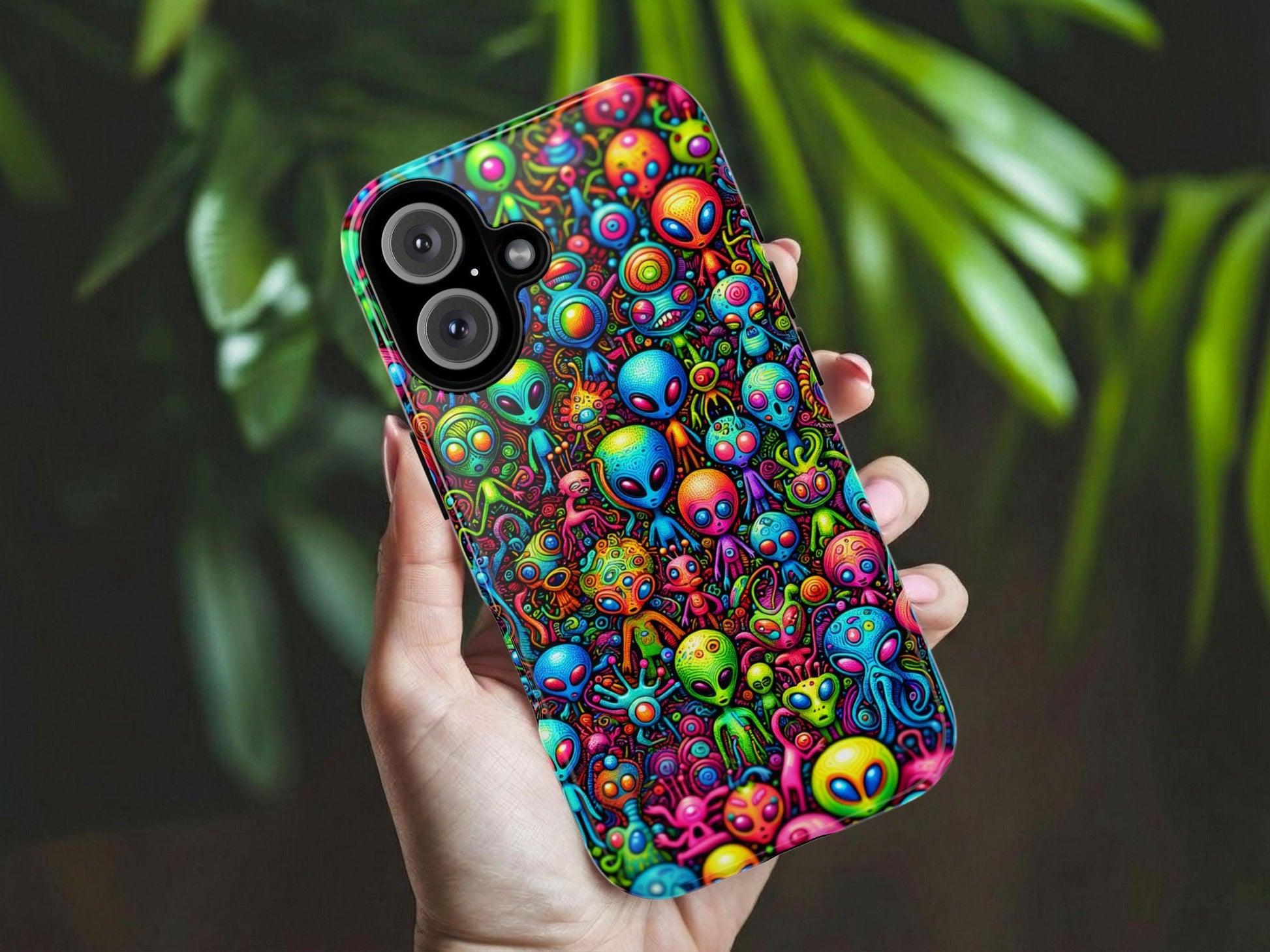 A hand holds the Whimsical Alien Phone Case by Golden Value SG features a vibrant collage of cartoon-style aliens and space creatures with unique shapes, big eyes, and vivid blue, pink, green, orange, and yellow hues densely packed together. Green foliage is softly blurred in the background.