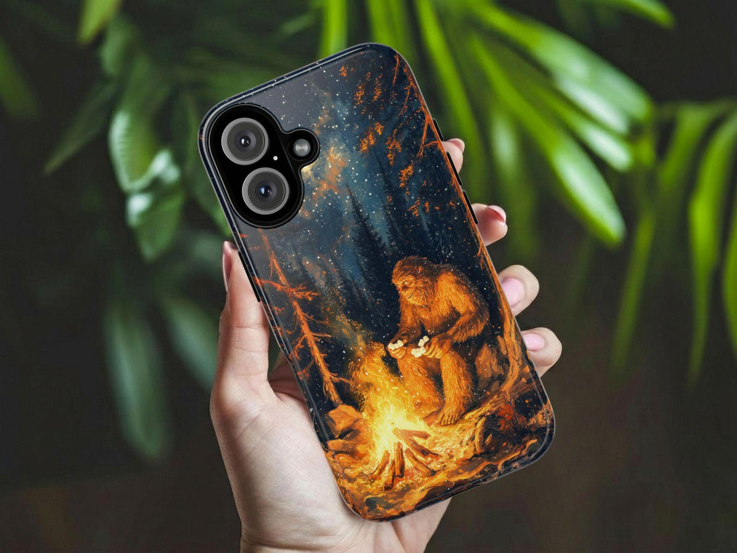 A hand holds the Bigfoot Phone Case by Golden Value SG featuring a cozy illustration of Bigfoot roasting marshmallows over a campfire at night, with a starry sky visible in the background. Green foliage is softly blurred in the background.