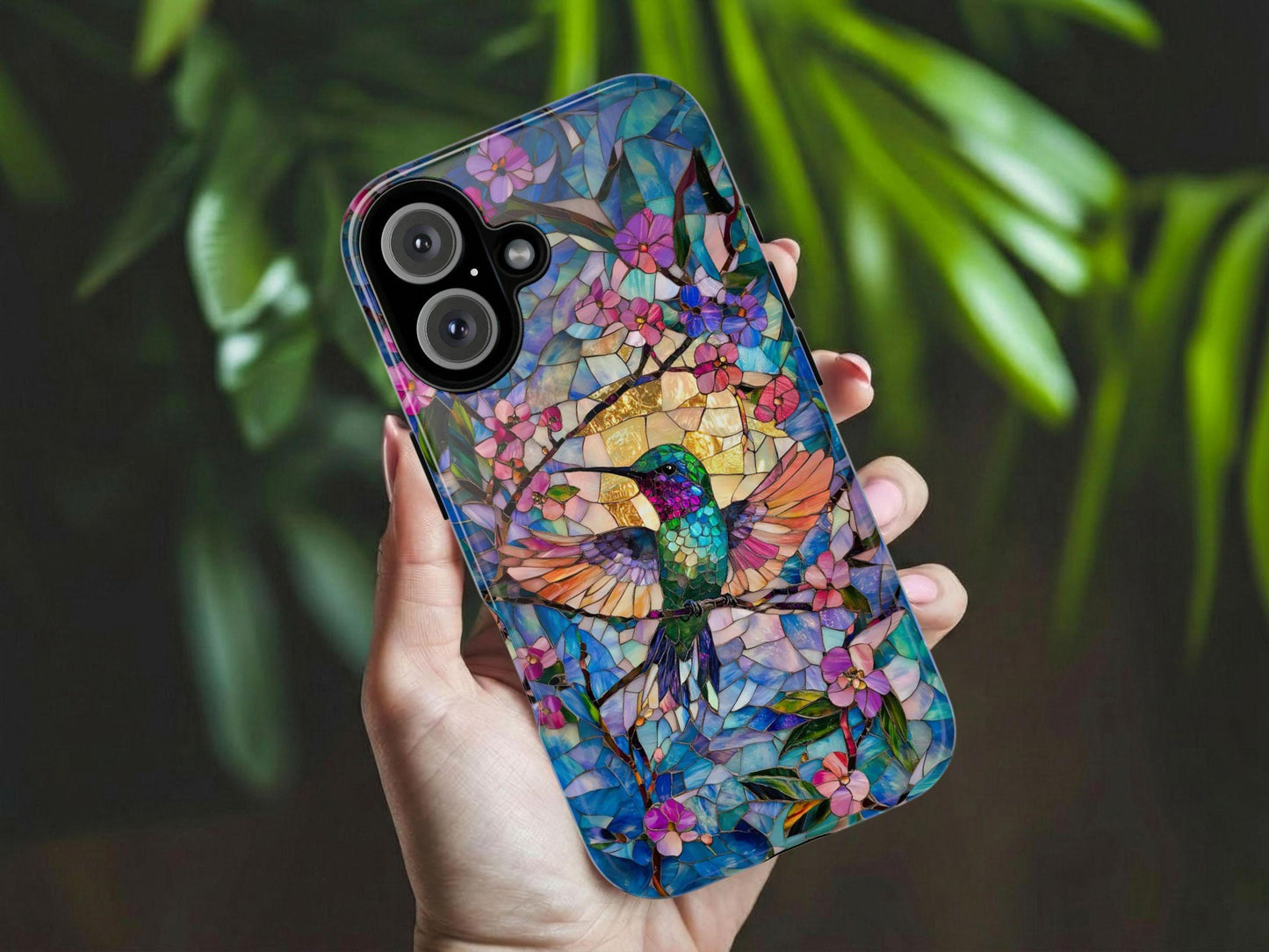 A hand holds the Hummingbird Phone Case by Golden Value SG features a colorful stained glass design with a vibrant hummingbird, pink flowers, green leaves, and a blue and gold background. Green foliage is softly blurred in the background.