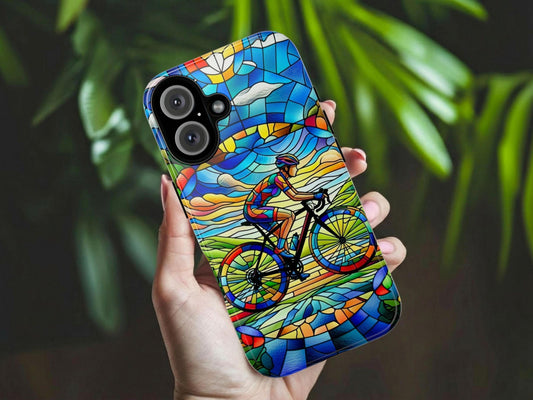 A hand holds a GOLDEN VALUE SG Female Cyclist Cycling Phone Case, featuring a stained glass-style design of a female cyclist riding through a vivid landscape with blue sky, clouds, and green hills. Blurred green leaves appear in the background.