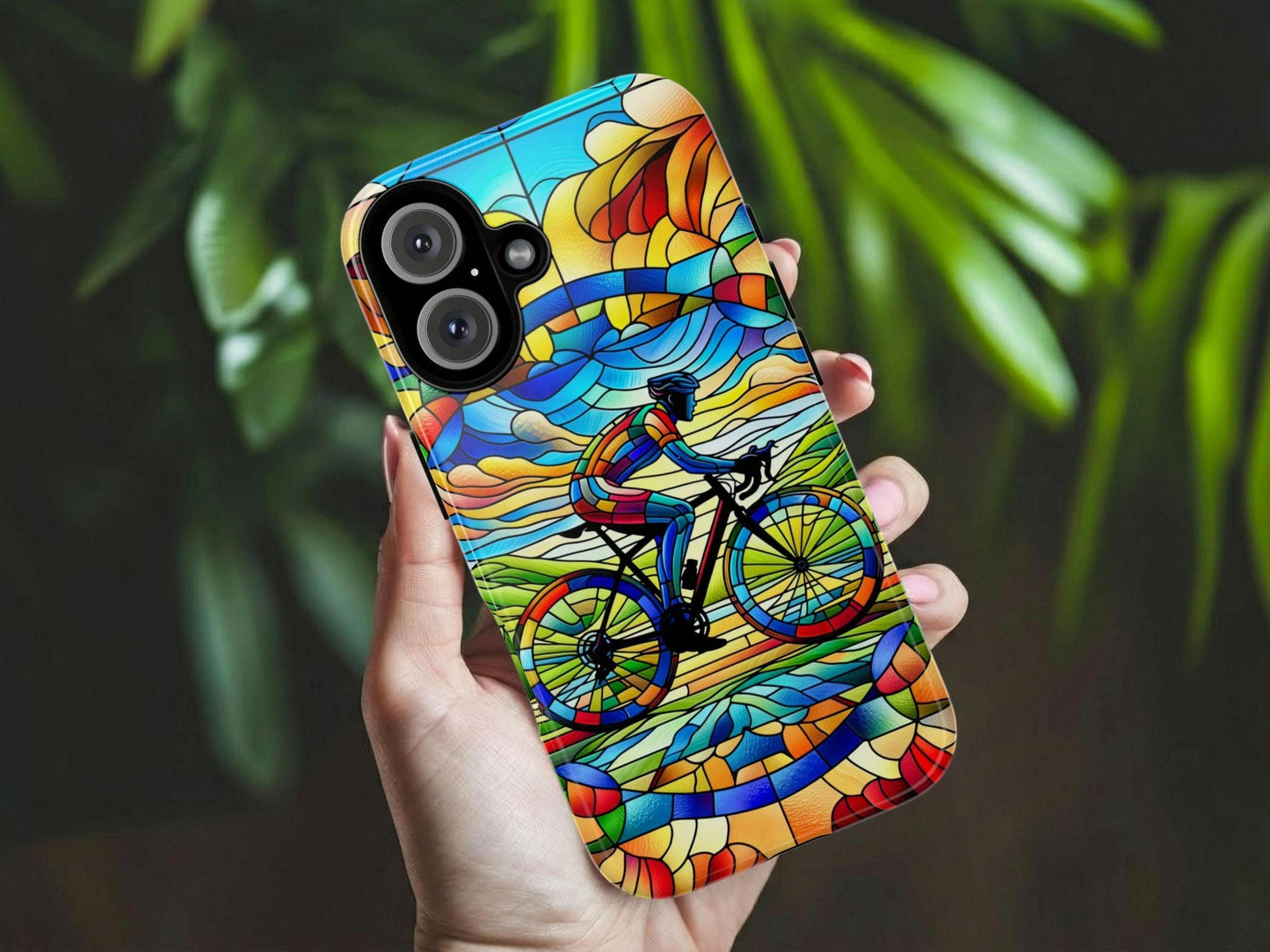 A hand holds the GOLDEN VALUE SG Cycling Phone Case, featuring a stained-glass style cyclist riding through a vibrant, abstract landscape in blue, green, yellow, and red. Blurred green plant leaves appear in the background.