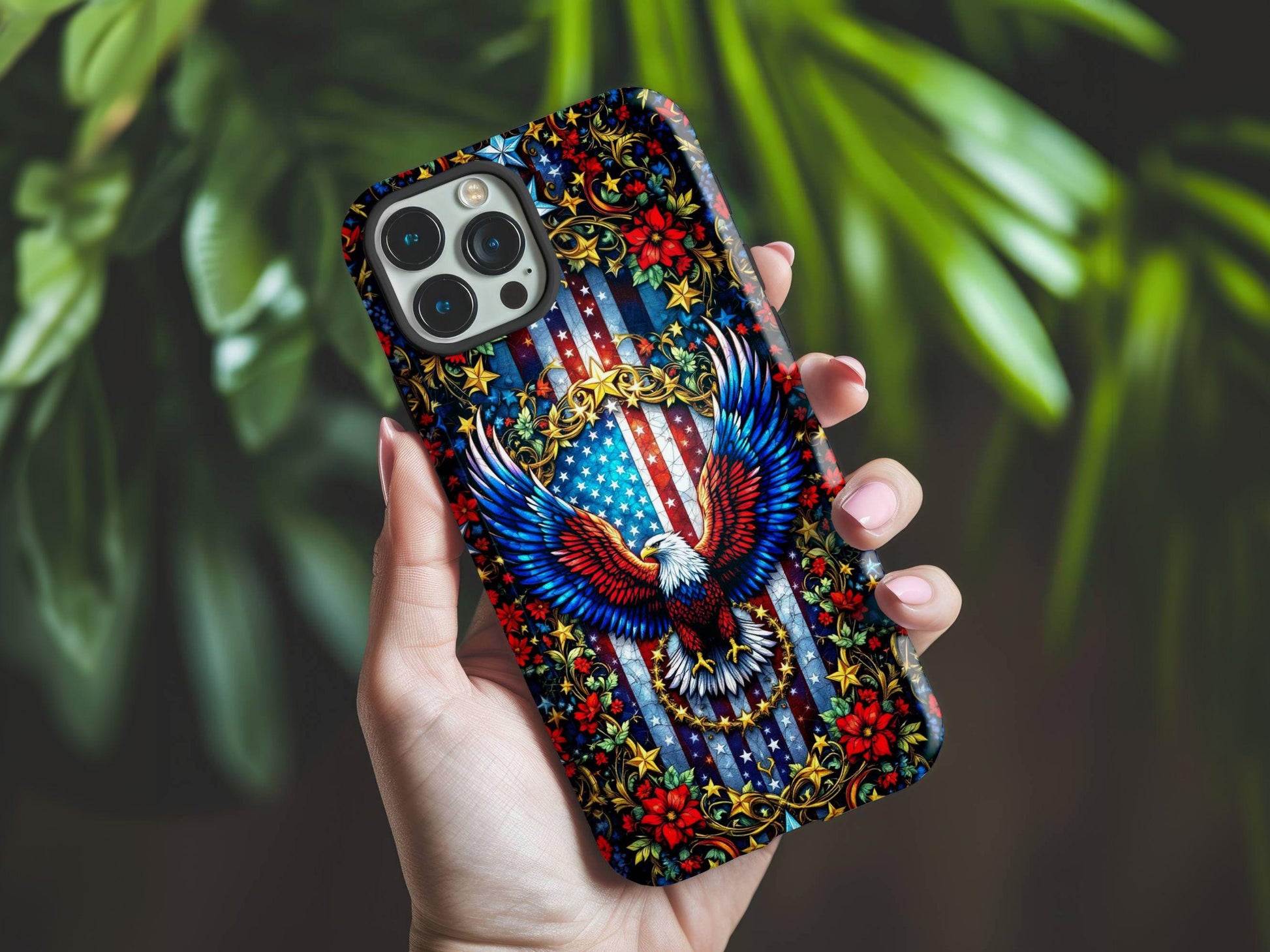 A hand holds the Golden Value SG American Flag and Eagle Phone Case features a vibrant stained glass-style bald eagle with spread wings, set against an American flag background with gold patterns, stars, and red flowers. Green foliage is softly blurred in the background.