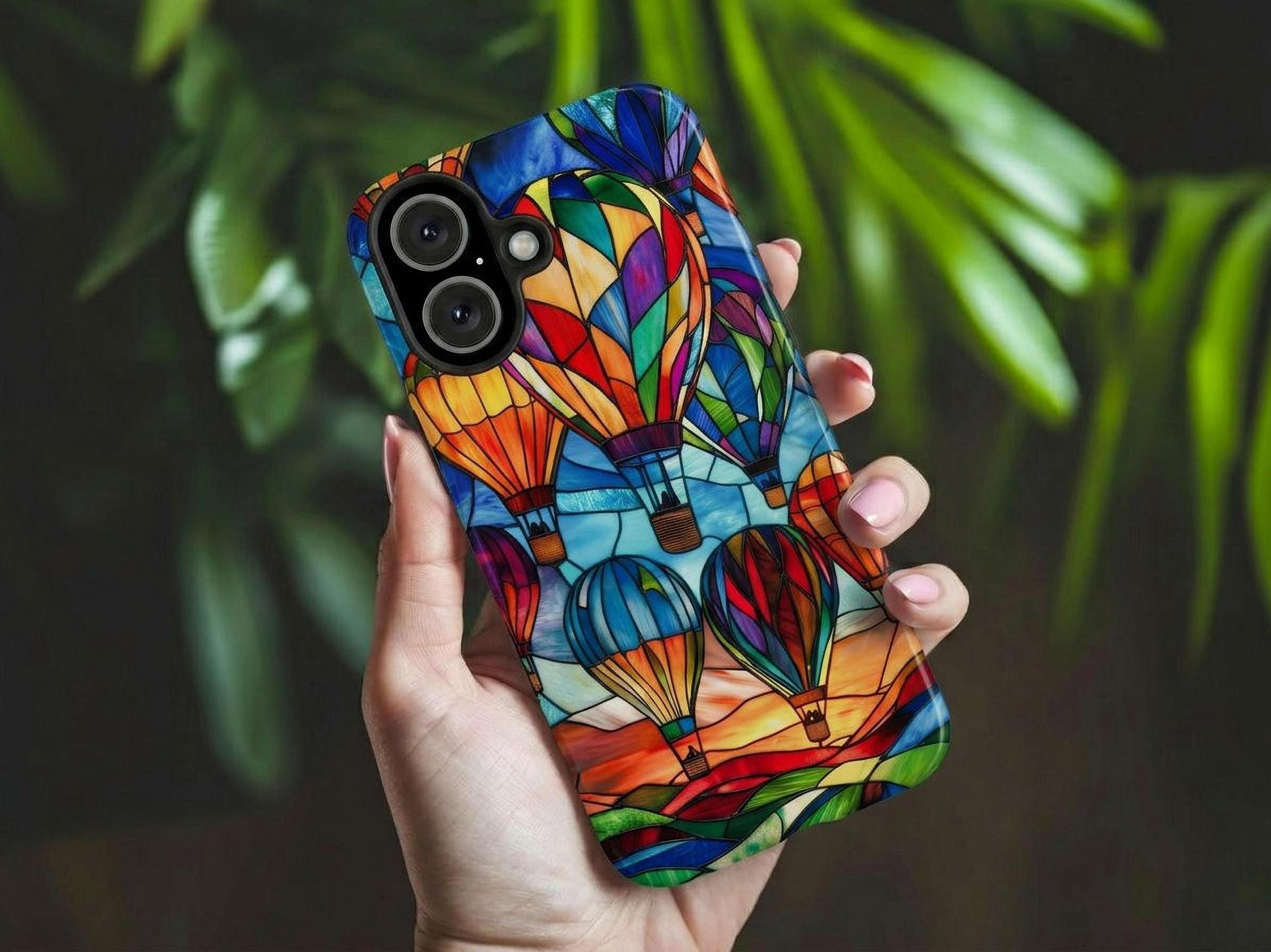 A hand holds the Golden Value SG's Hot Air Balloon Phone Case features stained glass artwork of vibrant hot air balloons in red, blue, green, and yellow against a sunset landscape with a sky turning from deep blue to orange. Green foliage is softly blurred in the background.
