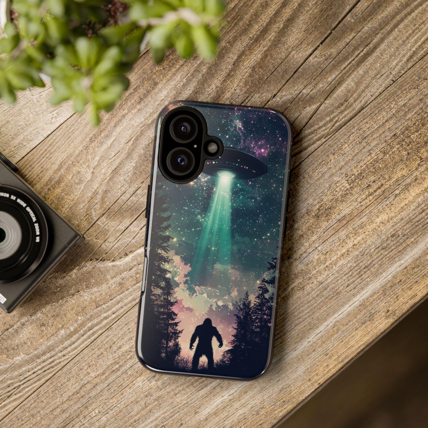 A Bigfoot UFO Phone Case by Golden Value SG featuring a night sky scene with a UFO and a silhouette of a Bigfoot-like creature. The case has a dark blue and purple galaxy design with a bright green light beam emanating from the UFO. The trees are silhouetted in black. On a wooden textured table.