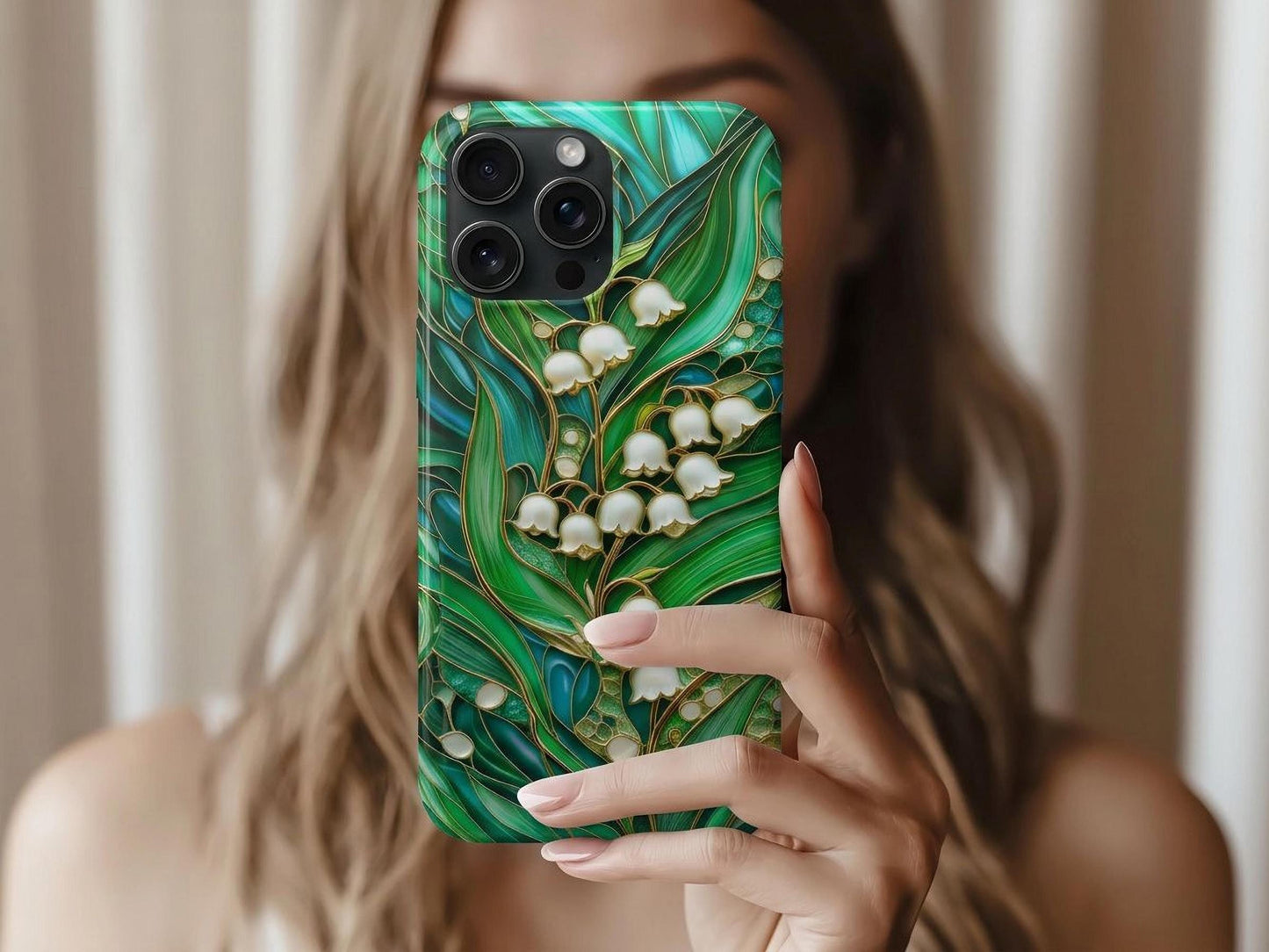 A woman with light brown hair holds up a Stained Glass Lily of the Valley Phone Case, showcasing a cluster of white lily of the valley flowers with golden stems and outlines, surrounded by lush green and blue-green stained glass leaves. The intricate design evokes natural beauty and sophisticated artistry.