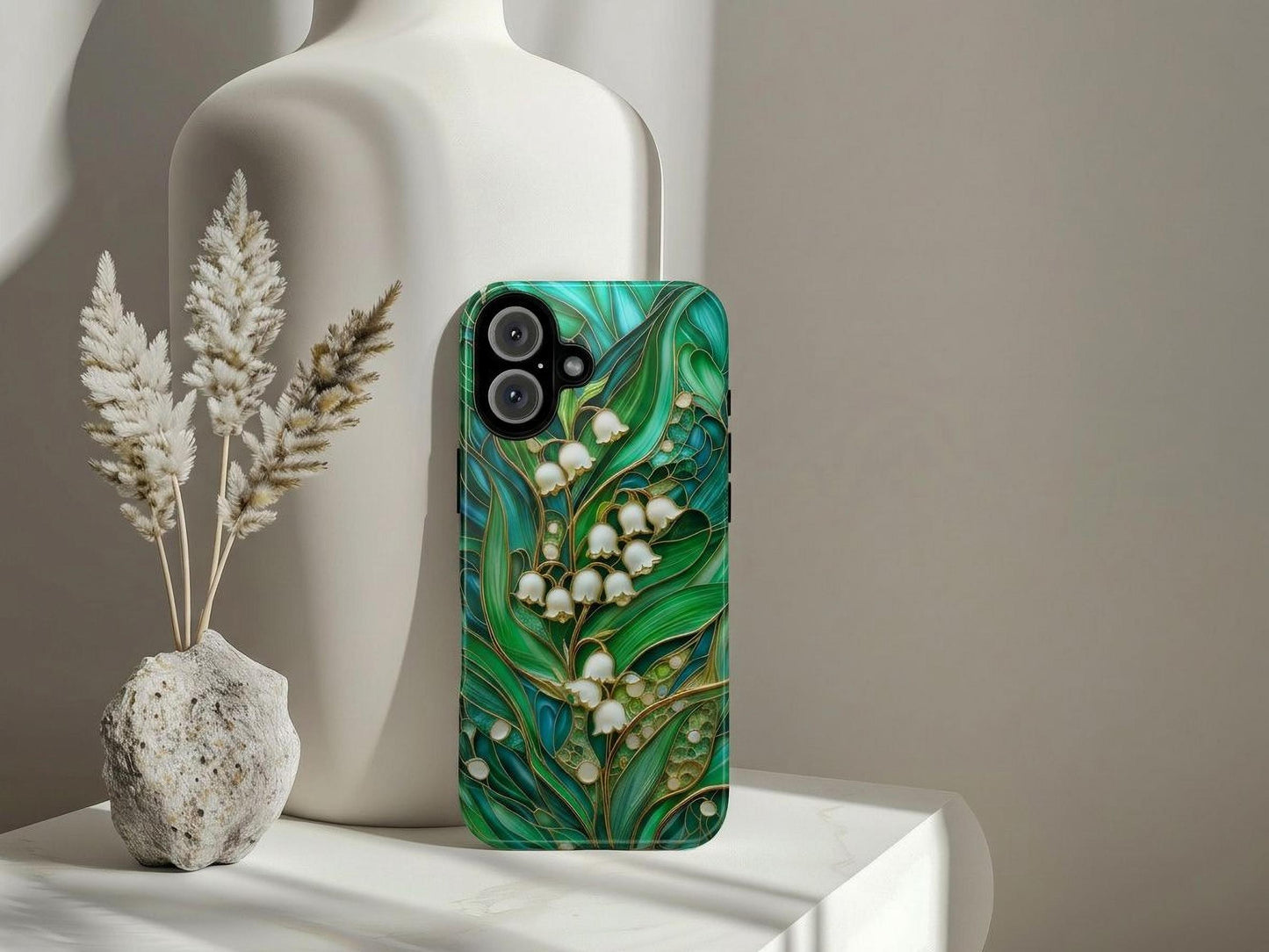 The Golden Value SG Stained Glass Lily of the Valley Phone Case, featuring a green leafy floral design with white lilies, stands upright by a white ceramic vase, with a small stone vase and beige dried grasses nearby on a softly lit white surface.