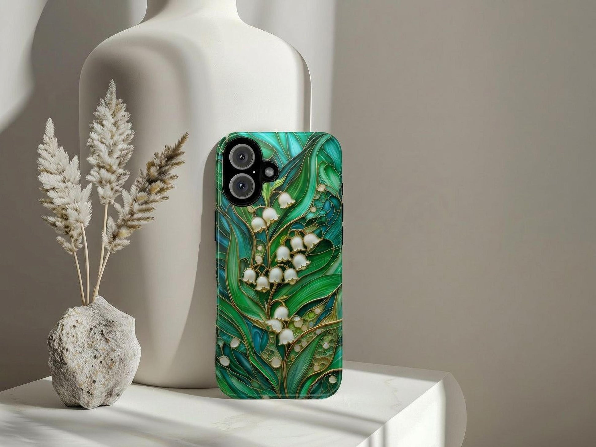 The Golden Value SG Stained Glass Lily of the Valley Phone Case, featuring a green leafy floral design with white lilies, stands upright by a white ceramic vase, with a small stone vase and beige dried grasses nearby on a softly lit white surface.