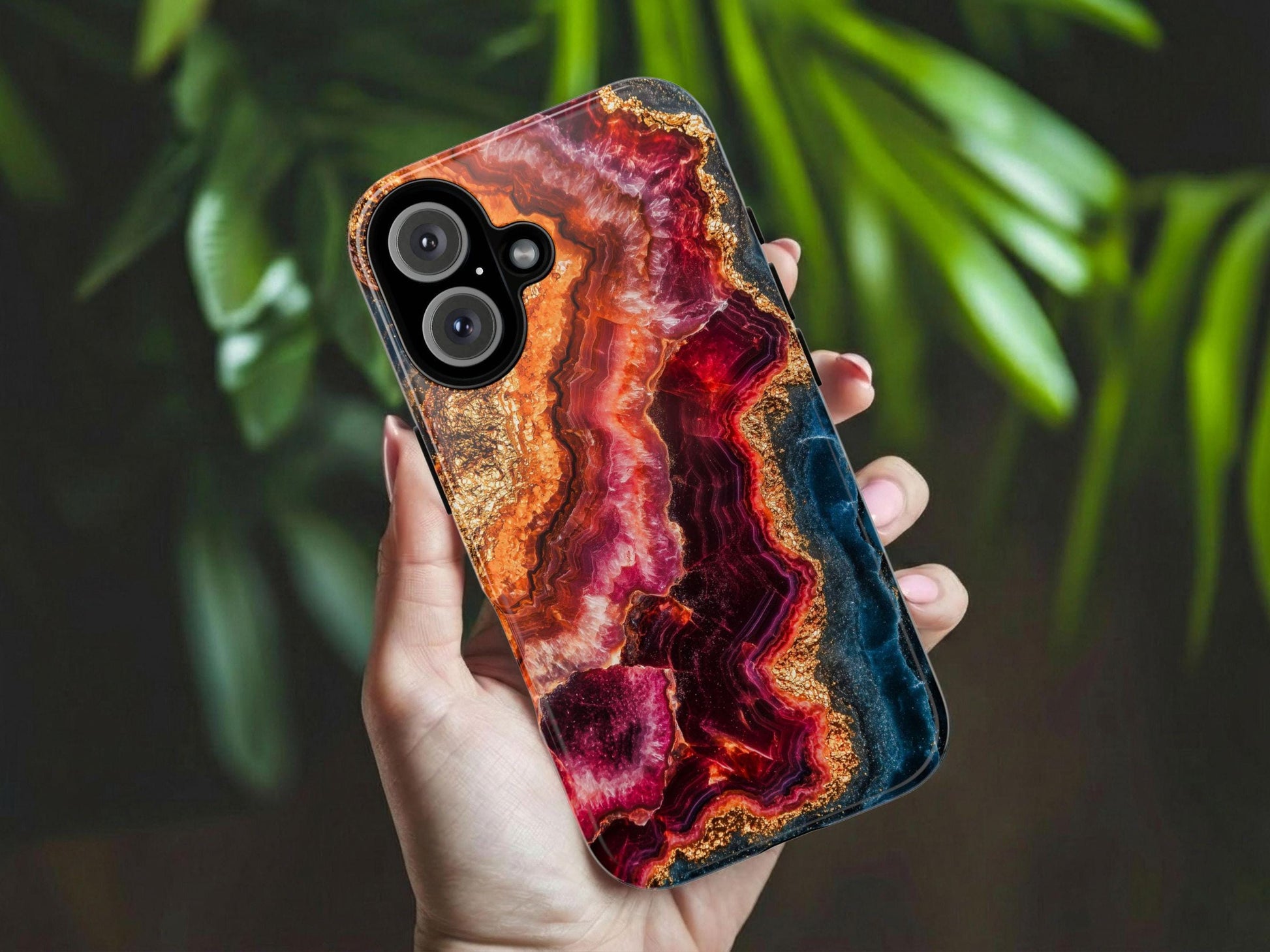 A hand holds the Elegant Ruby Crystal Phone Case by Golden Value SG, displaying a vibrant, abstract agate-inspired pattern in red, orange, gold, and black. Blurred green leaves are visible in the background.
