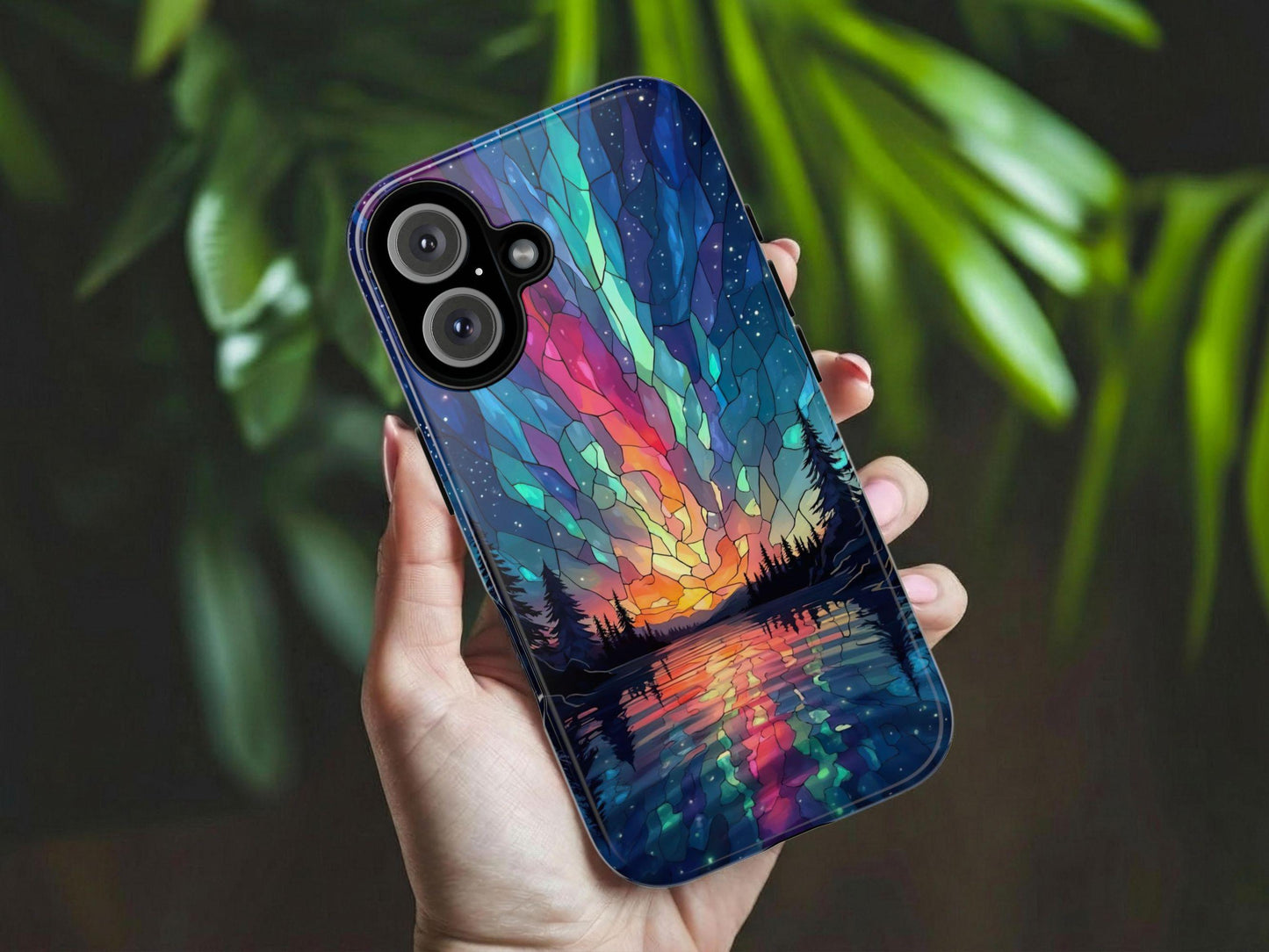 A hand holds the Golden Value SG Aurora Inspired Design Stained Glass Phone Case, featuring a colorful sunset landscape with trees and a lake under a starry sky. Blurred green leaves appear in the background.