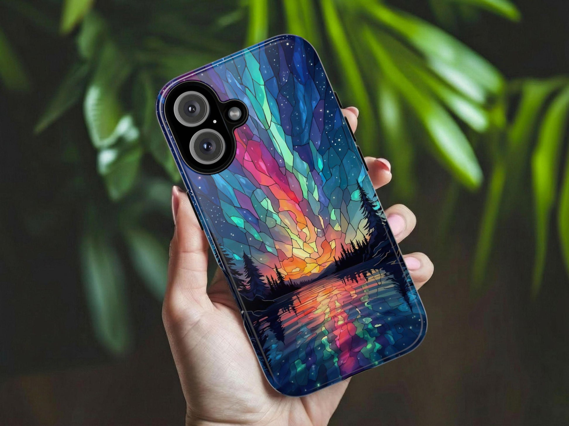 A hand holds the Golden Value SG Aurora Inspired Design Stained Glass Phone Case, featuring a colorful sunset landscape with trees and a lake under a starry sky. Blurred green leaves appear in the background.