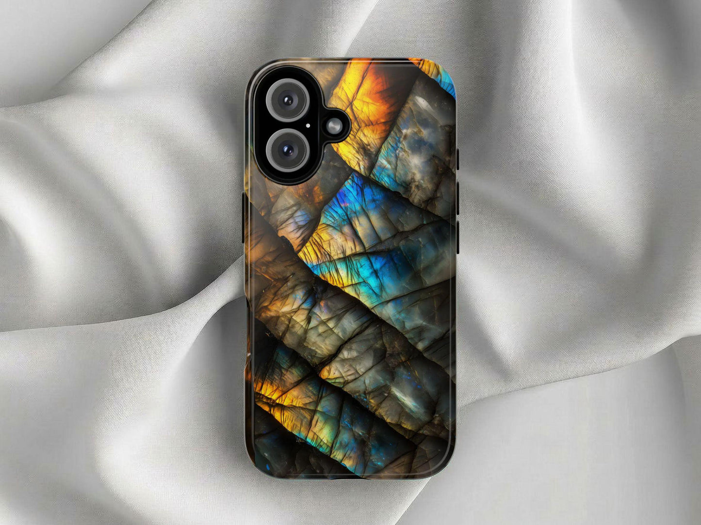 The Golden Value SG Labradorite Gemstone Phone Case, with its abstract iridescent blue, gold, and orange design, lies on softly draped light gray satin fabric.