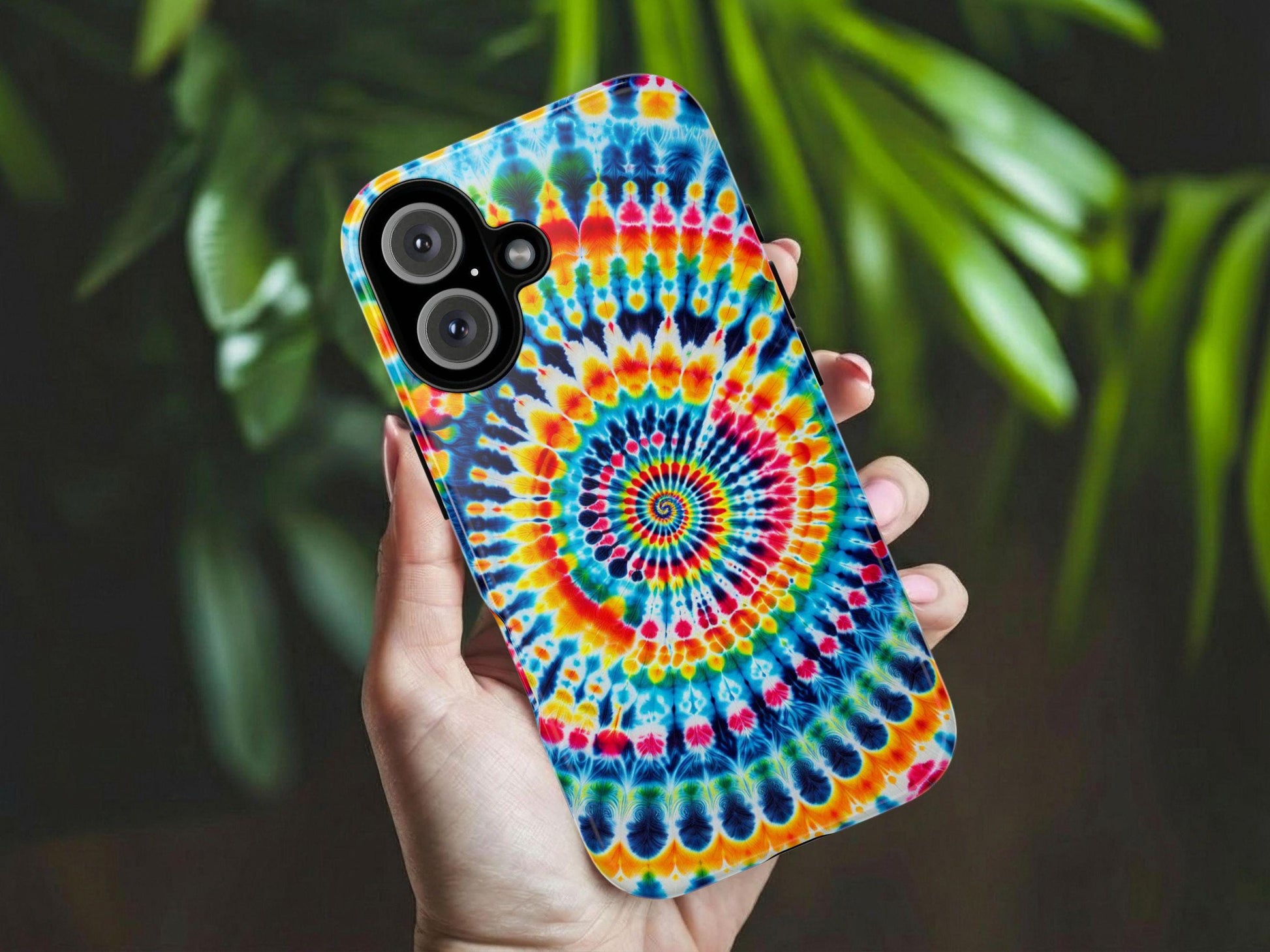 A hand holds the Golden Value SG Tie Dye Phone Case features a vibrant spiral design in yellow, red, blue, and teal, radiating outward with symmetrical bursts and intricate shapes. Green foliage is softly blurred in the background.