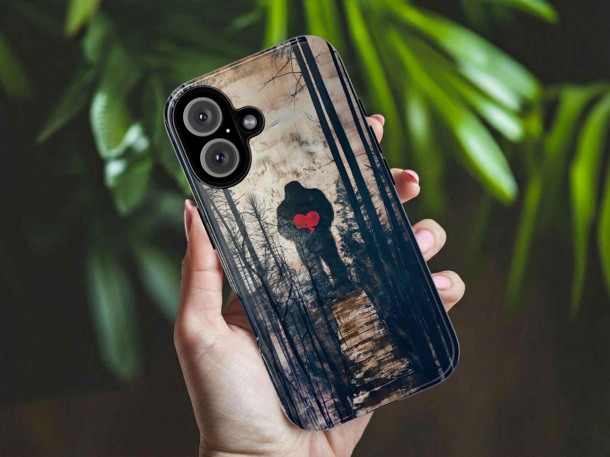 A hand holds the Golden Value SG Bigfoot Silhouette Phone Case, showing Bigfoot in a forest holding a red heart. Green leaves blur softly in the background.