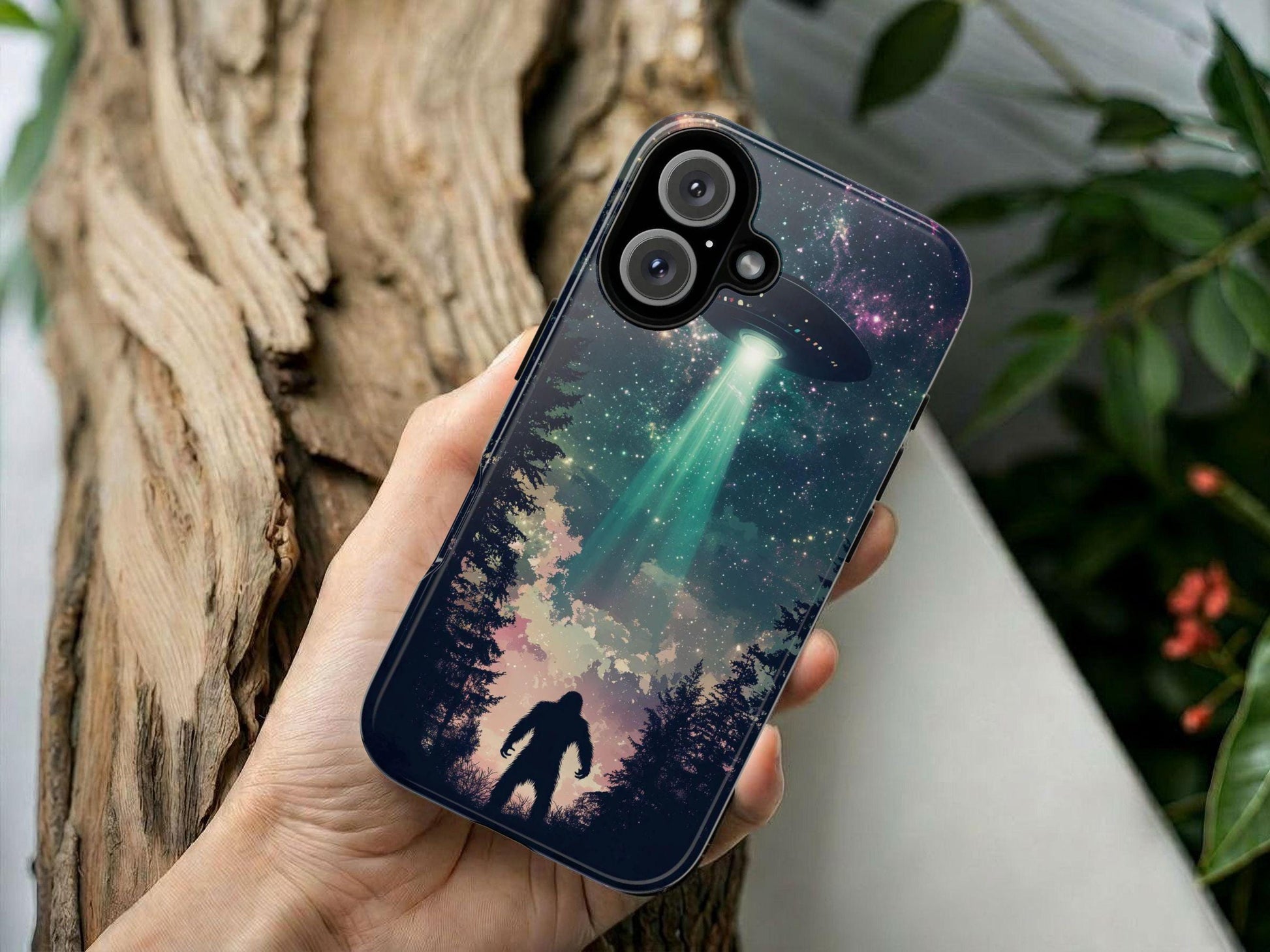 A hand holds the Golden Value SG Bigfoot UFO Phone Case, showing forest art at night with a Bigfoot silhouette and a UFO beaming green light, set against a blurred wood and leafy background.