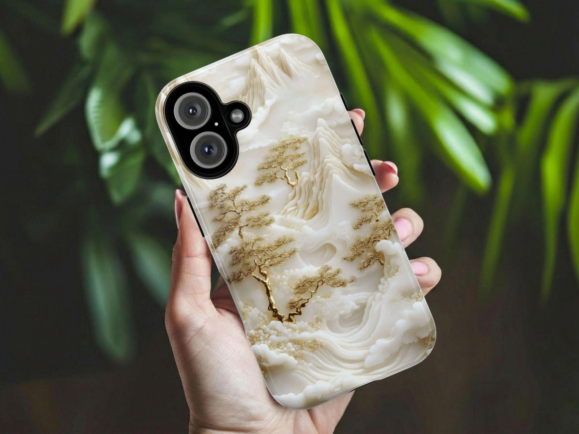 A hand holds the Unique White Jade Carving Phone Case by Golden Value SG features an intricately carved design of mountains, clouds, and golden pine trees in a monochrome palette, creating a textured, ethereal, and serene atmosphere. Green foliage is softly blurred in the background.