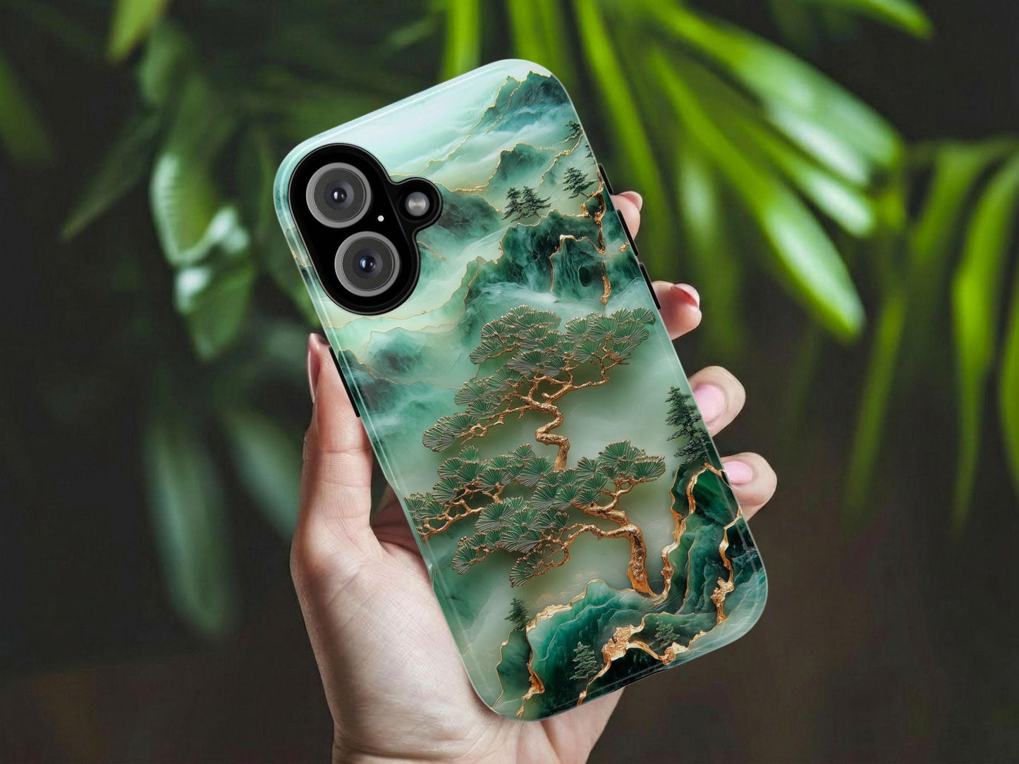 A hand with light pink nails holds the Unique Jade Carving Phone Case by Golden Value SG, featuring a gold-embellished tree and misty mountain scene; leafy plants are softly blurred in the background.