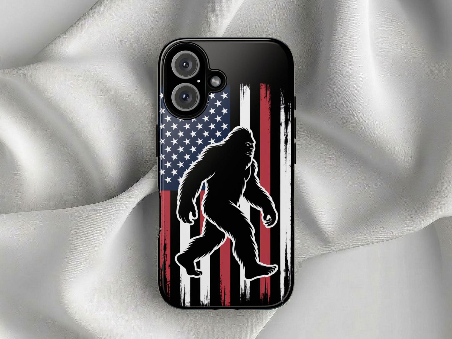 The Golden Value SG Bigfoot Silhouette Phone Case features a bold Bigfoot outline before a distressed American flag on a glossy black surface, displayed on light, crumpled fabric.
