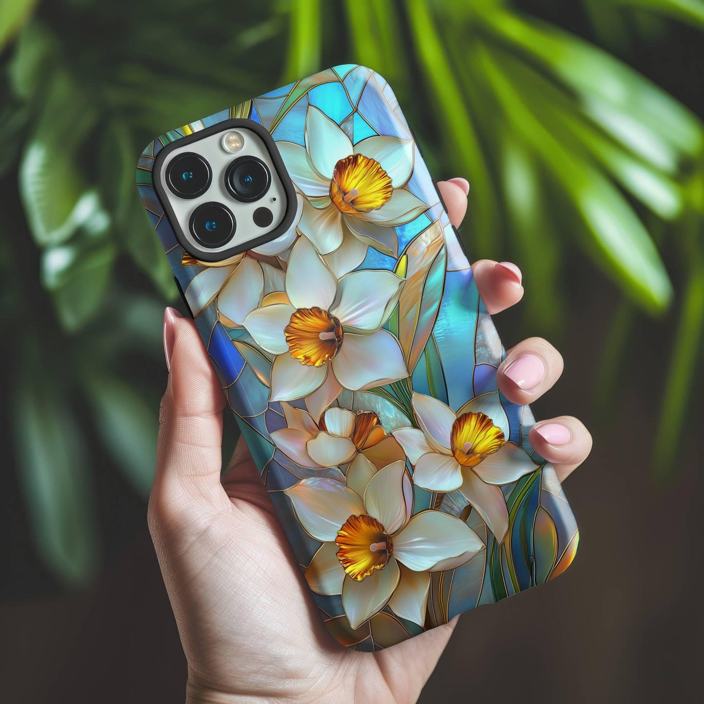 A hand holds a Stained Glass Daffodil Phone Case by Golden Value SG featuring elegant white daffodils with vibrant yellow centers and green leaves against a mosaic-style blue and teal background. This floral design captures the delicate beauty of spring flowers in a colorful, art-inspired pattern, perfect for adding a touch of artistic charm to your device. Green foliage is softly blurred in the background.