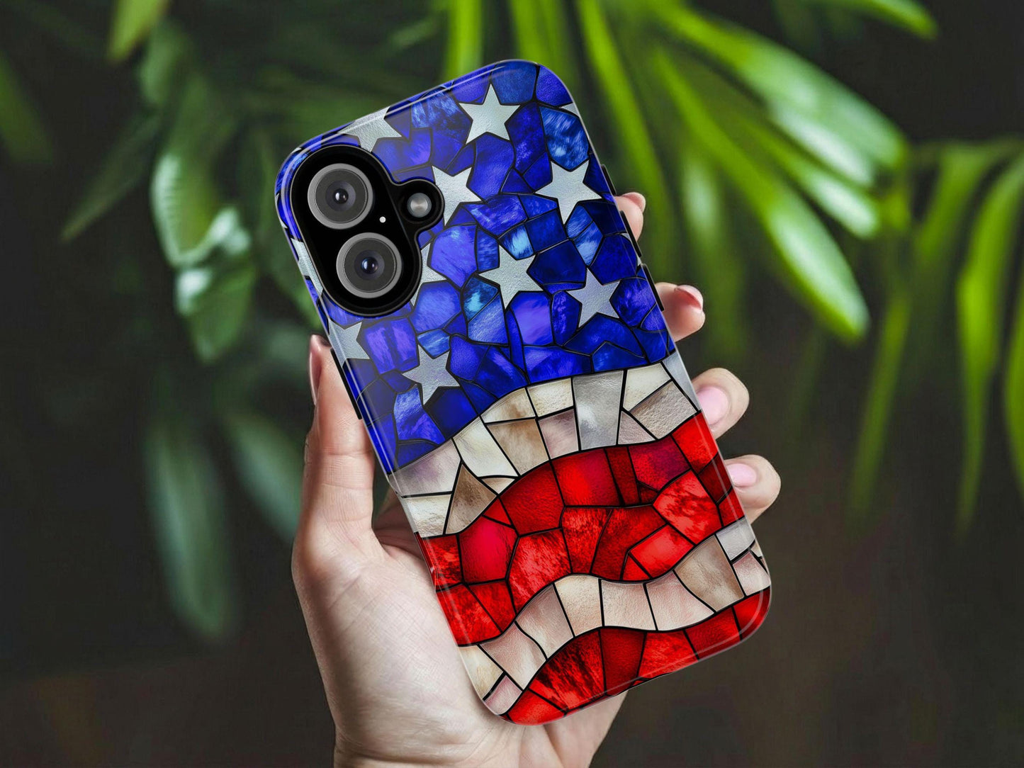 A hand holds the Golden Value SG American Flag Phone Case, featuring a mosaic design with blue and white stars at the top and red and white stripes below. Blurred green leaves appear in the background.