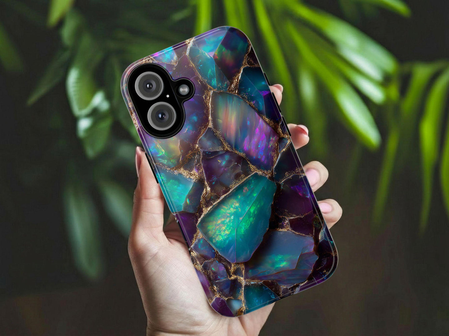 A hand holds the Vibrant Iridescent Opal Phone Case by Golden Value SG featuring a mosaic of deep purple, teal blue, and green gemstones with golden outlines. Captures a mystical, luxurious, and shimmering raw crystal aesthetic, offering a unique and eye-catching accessory. Green foliage is softly blurred in the background.