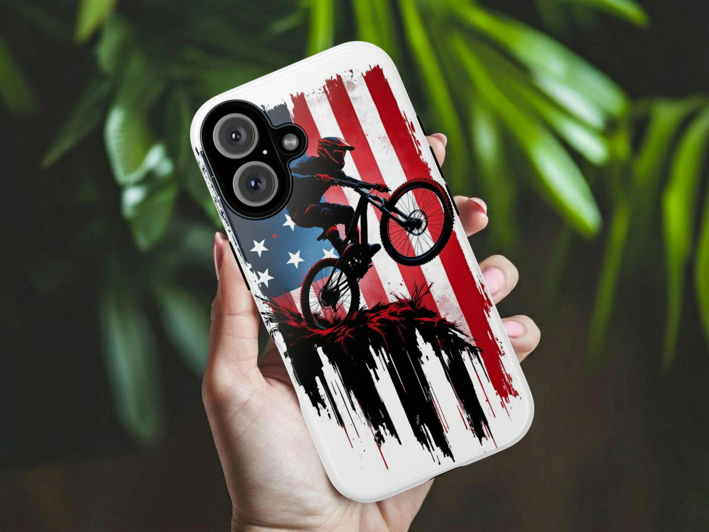 A hand holds the Mountain Biker Phone Case by Golden Value SG featuring a dark silhouette of a mountain biker mid-jump against a distressed, grunge-style American flag backdrop in red, white, and blue. The bold graphic is accented with dramatic black ink drip effects and red splatter details at the base, creating a powerful, patriotic, action-sport aesthetic. Perfect for mountain biking enthusiasts and proud Americans. Green foliage is softly blurred in the background.