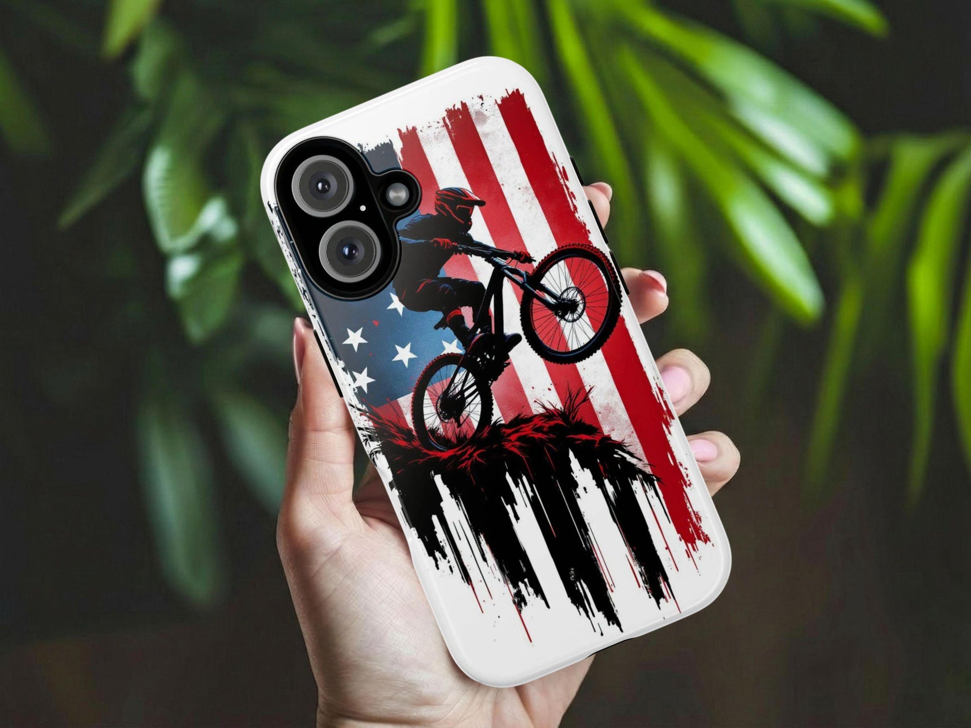 A hand holds the Mountain Biker Phone Case by Golden Value SG featuring a dark silhouette of a mountain biker mid-jump against a distressed, grunge-style American flag backdrop in red, white, and blue. The bold graphic is accented with dramatic black ink drip effects and red splatter details at the base, creating a powerful, patriotic, action-sport aesthetic. Perfect for mountain biking enthusiasts and proud Americans. Green foliage is softly blurred in the background.