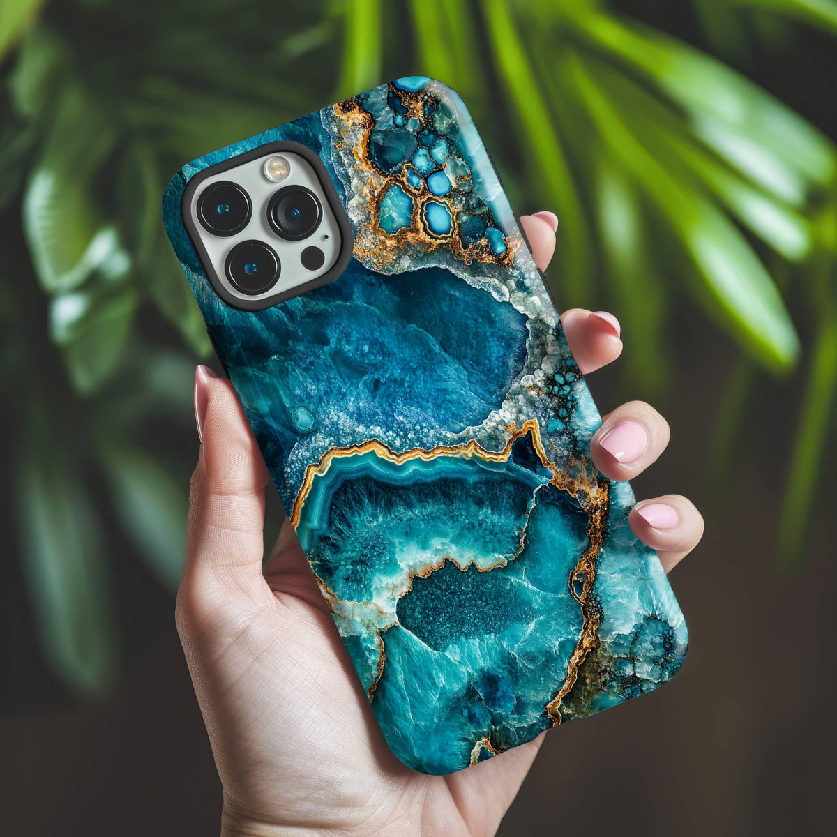 A hand holds the December Birthstone Turquoise Crystal Geode Design Phone Case by Golden Value SG featuring the intricate patterns of rich blue and teal hues evoke the enchanting allure of geological formations. Green foliage is softly blurred in the background.