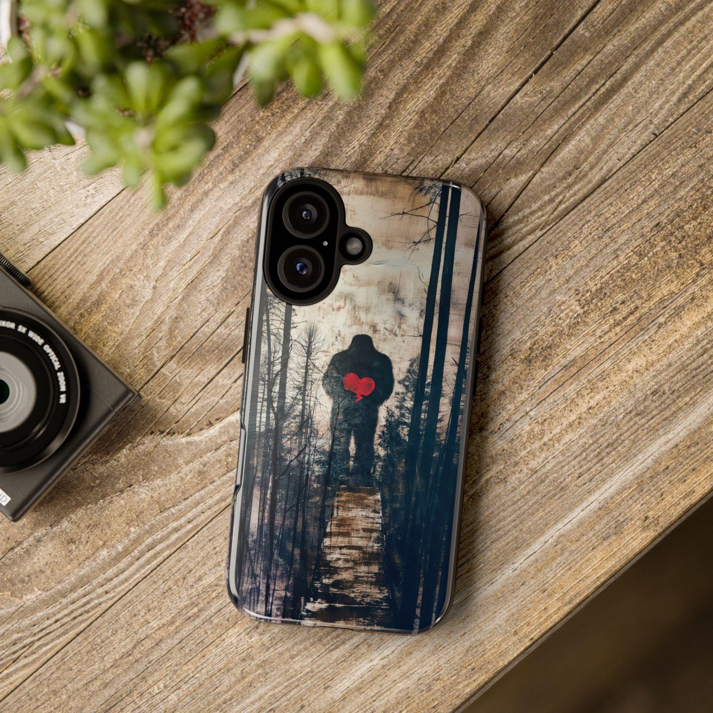 A phone case featuring a forest scene with Bigfoot holding a red heart sits on a wooden table next to a camera lens and green plant.