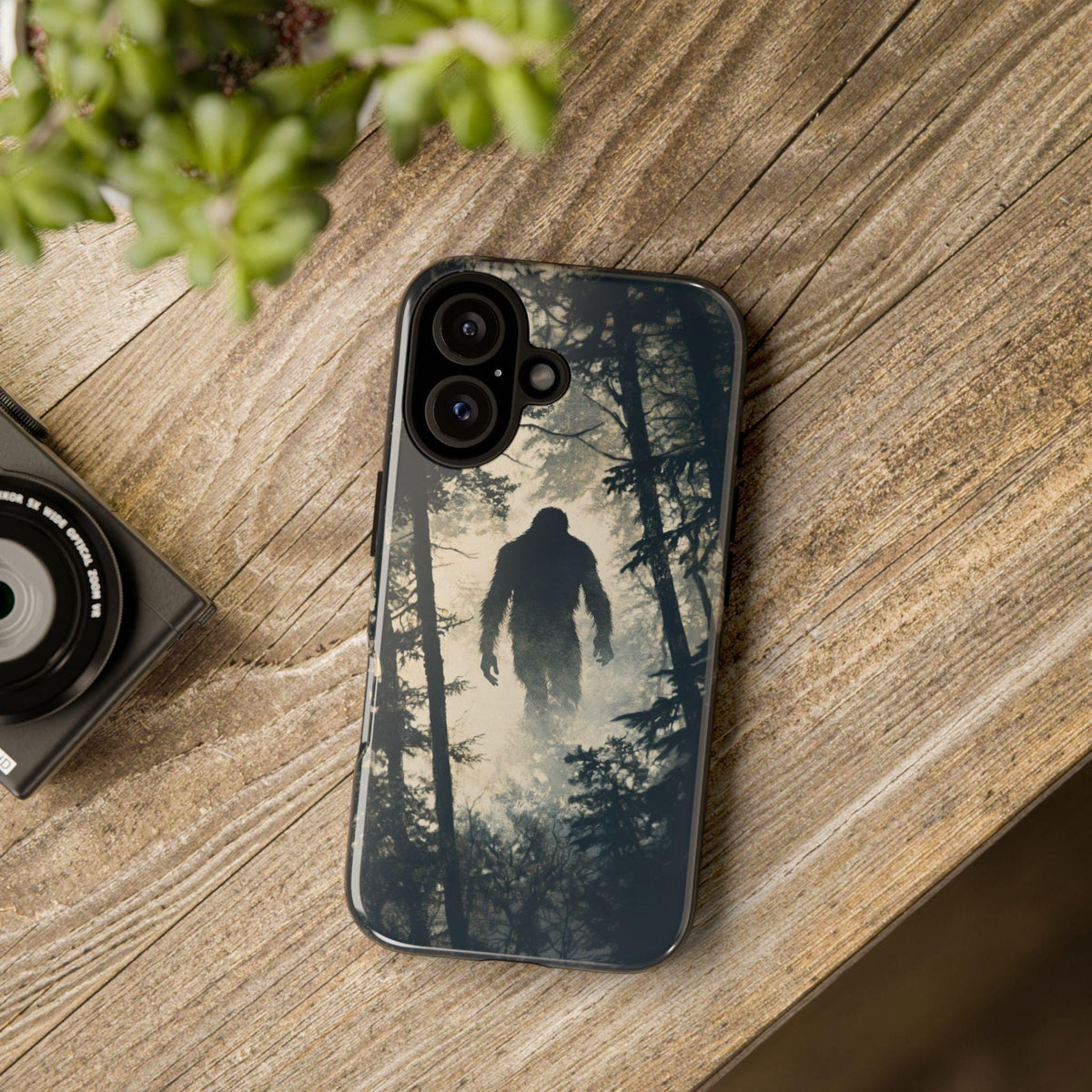 A smartphone case featuring a Bigfoot silhouette in a misty forest rests on a wooden surface next to a camera lens and a small potted plant.
