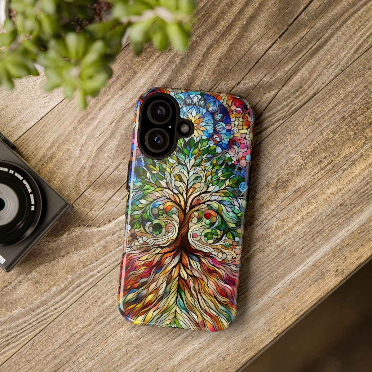 A smartphone case with a colorful stained-glass style tree of life design rests on a wooden surface next to a camera and green plant leaves.