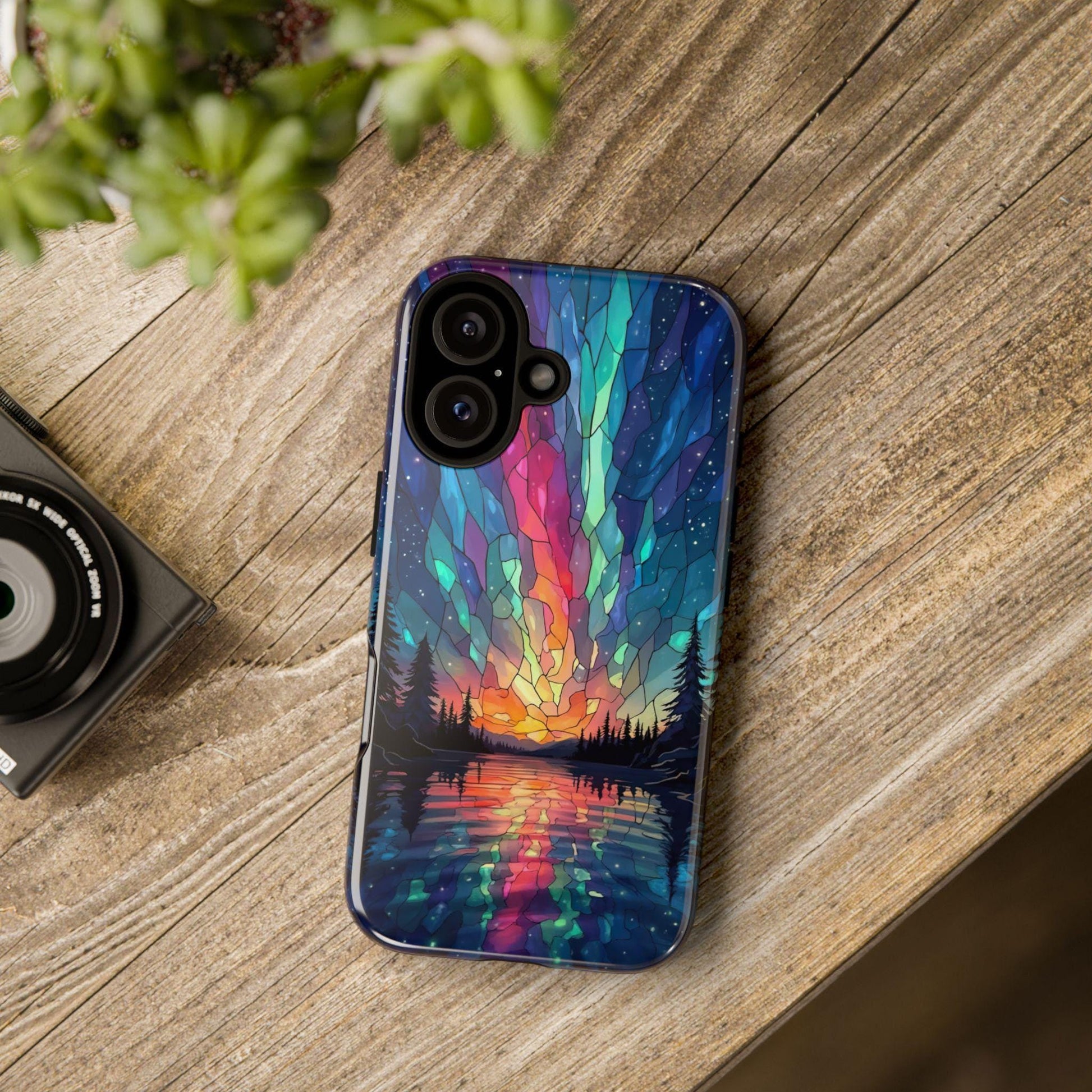 A phone case with a northern lights stained glass graphic lies on a wooden surface next to a camera lens and a plant.