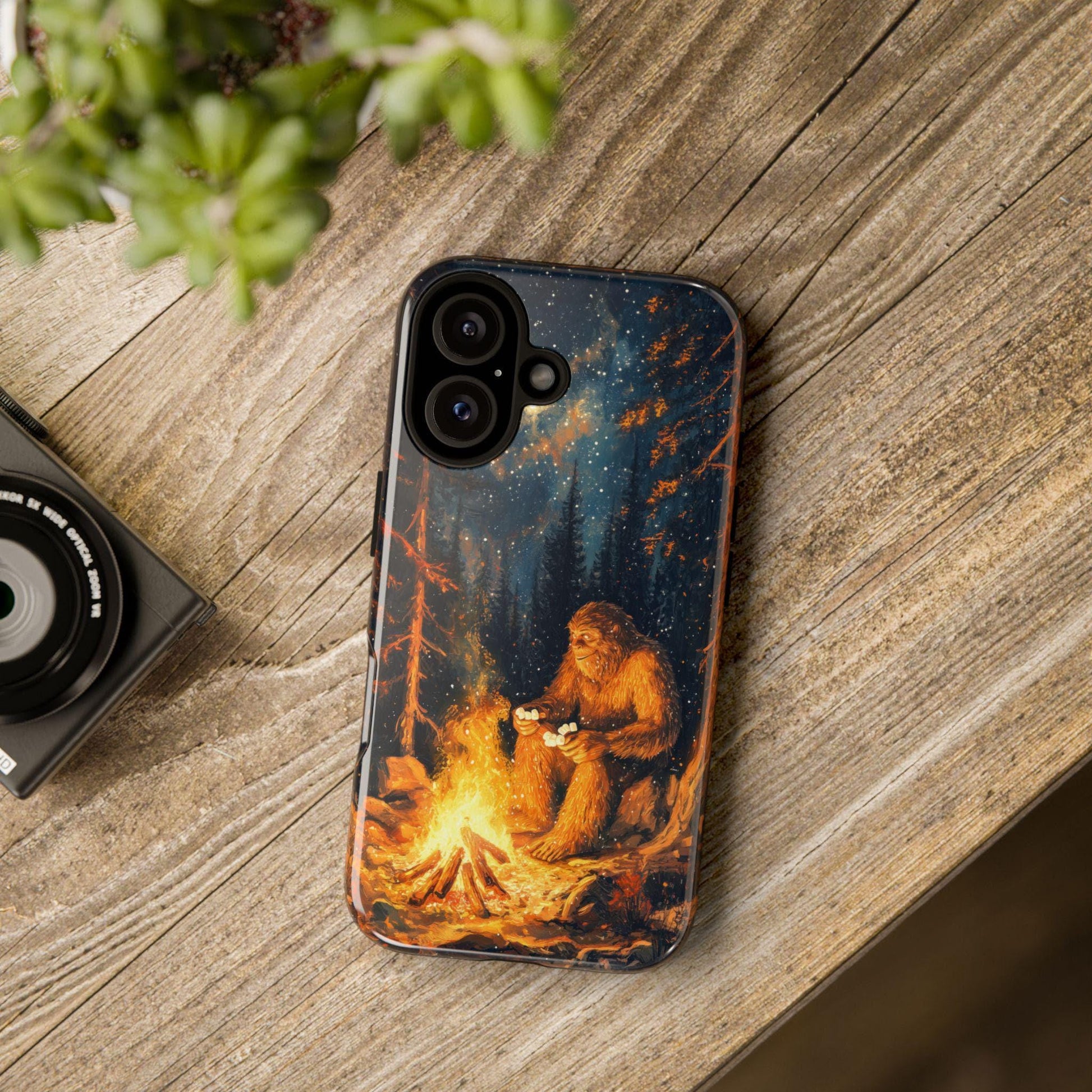 A smartphone case featuring a graphic of Bigfoot roasting marshmallows by a campfire in a forest under a starry night sky, resting on a wooden surface next to a camera lens and a small potted plant.