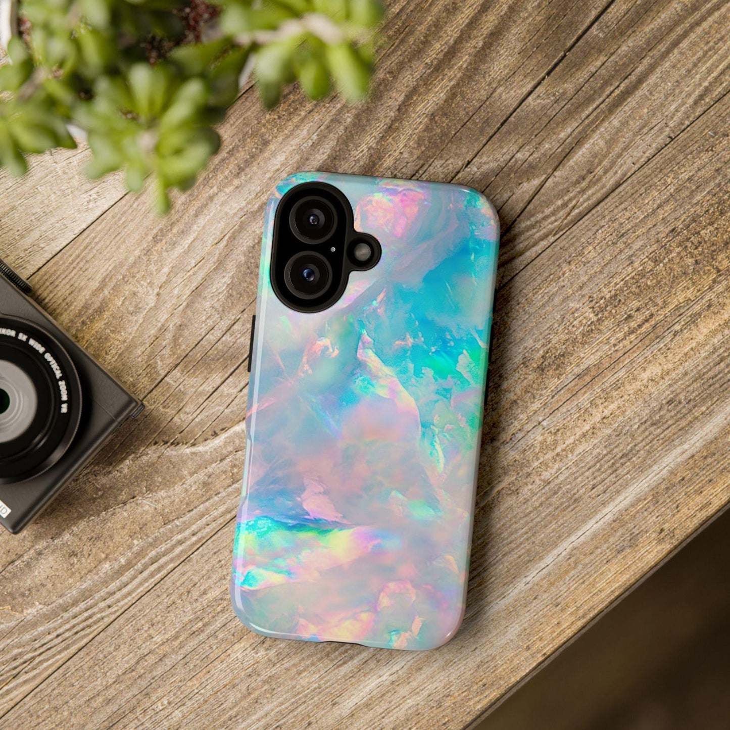 A phone case featuring a pastel iridescent opal pattern lies flat on a wooden surface next to a camera and green plant.