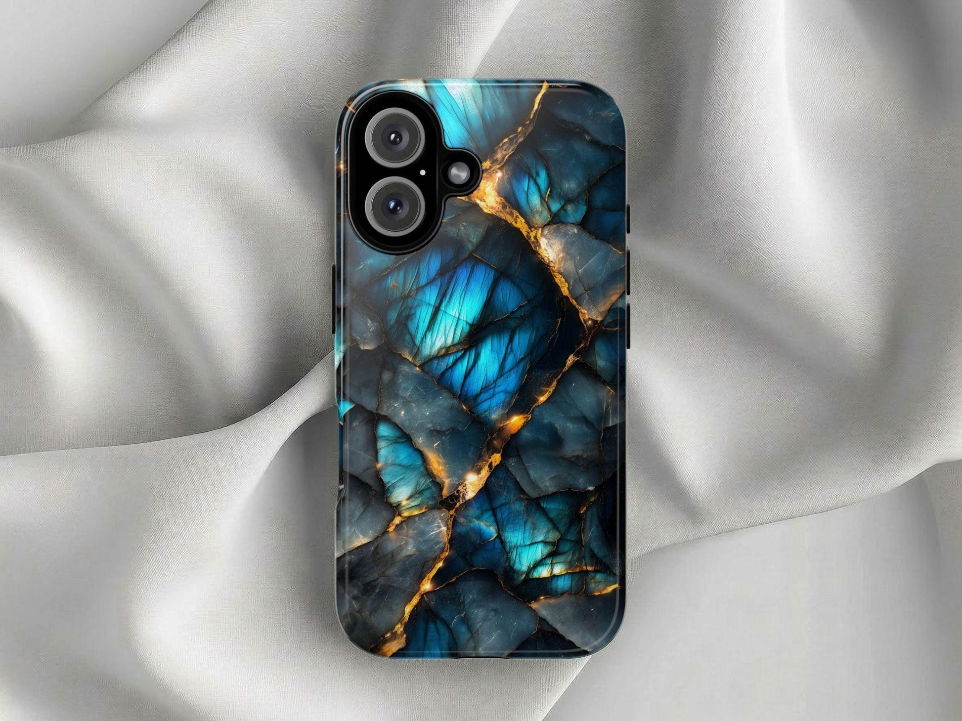 A smartphone case with a blue and black labradorite crystal pattern featuring gold veins lies on a white draped fabric.