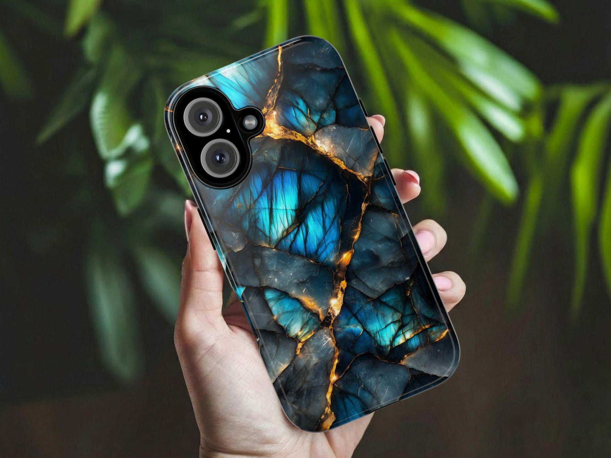A person holds up a labradorite gemstone patterned phone case against a blurred green plant background.