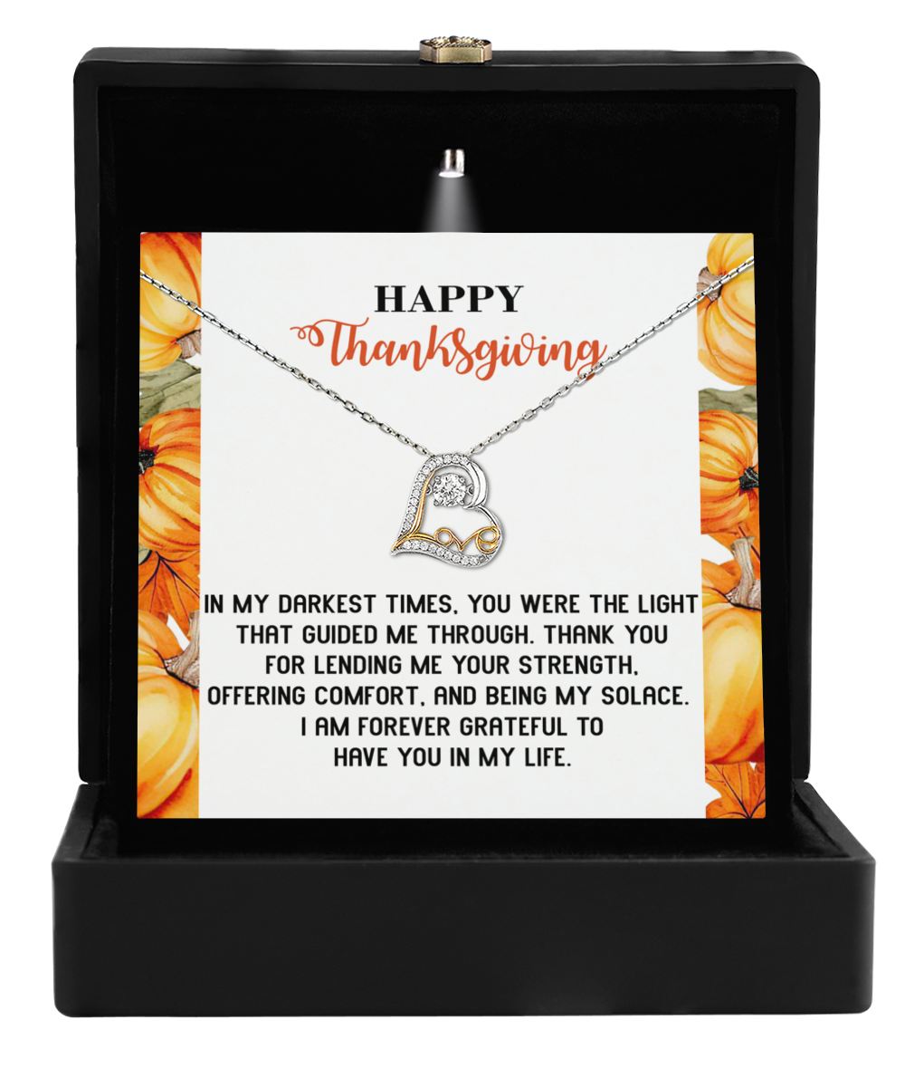 A Thanksgiving-Guided Me Through - Love Dancing Necklace, made of rhodium-plated silver and shaped like a heart pendant, rests in a black jewelry box. Behind it, a Thanksgiving card features pumpkin illustrations and a message of gratitude.